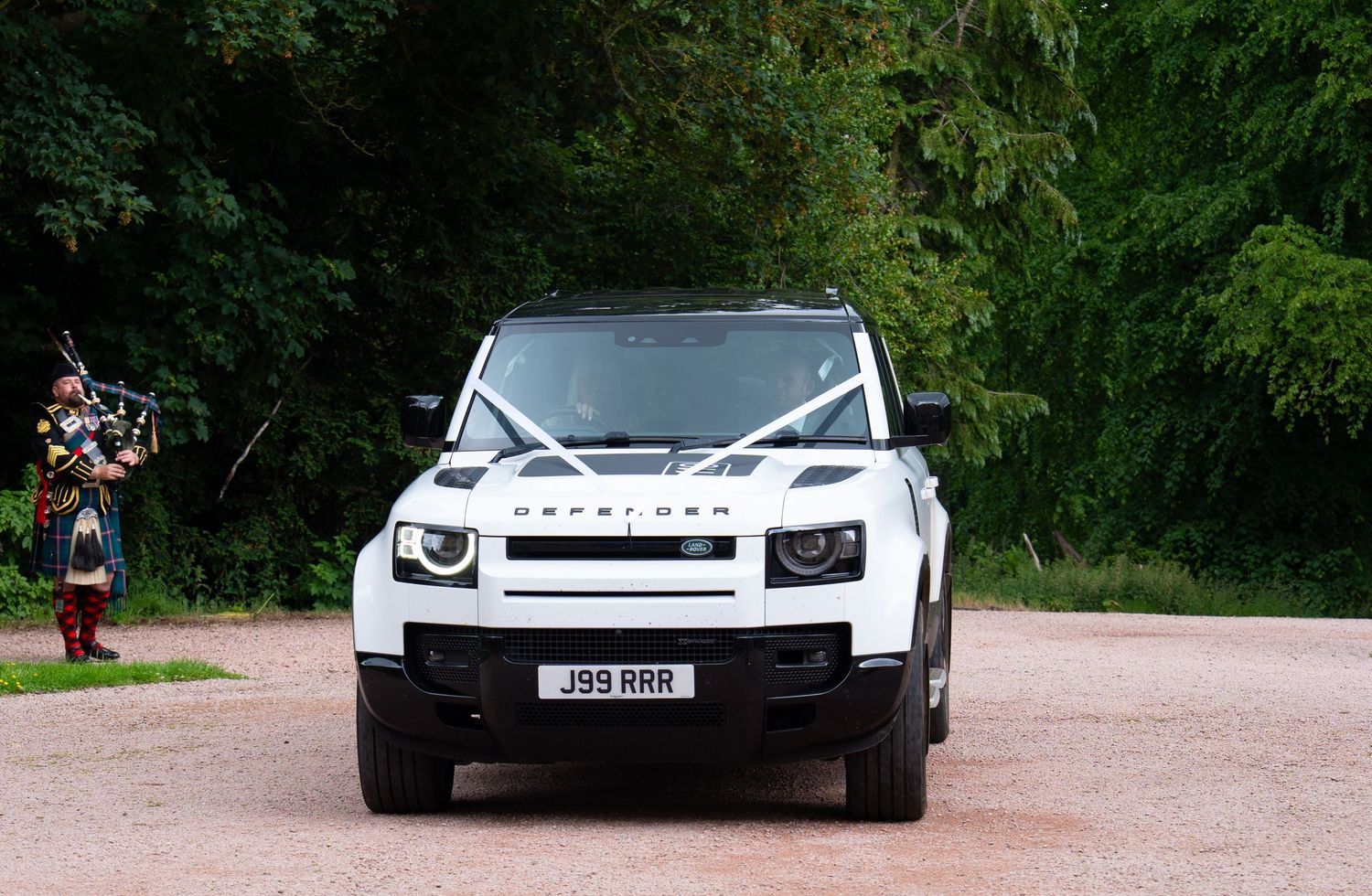 A line of luxury cars including Land Rovers and Range Rovers parked on a gravel driveway surrounded by trees.
