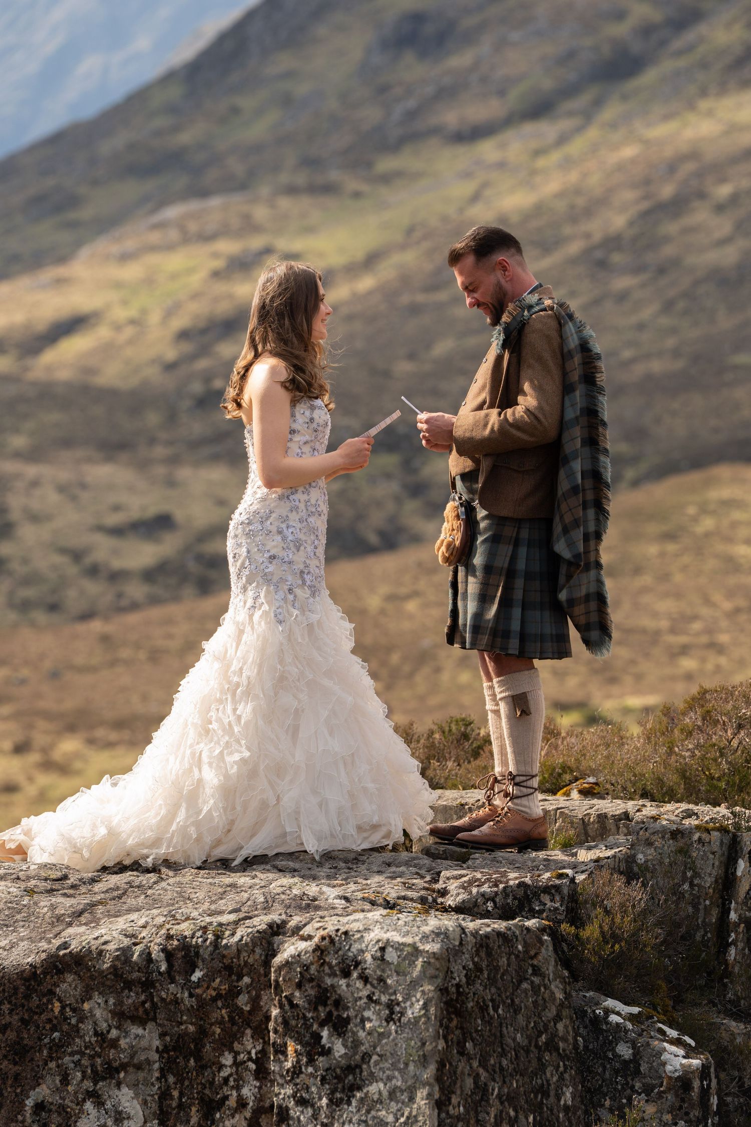 Couple in wedding attire exchanging vows on rocky outcrop in Scottish mountains.