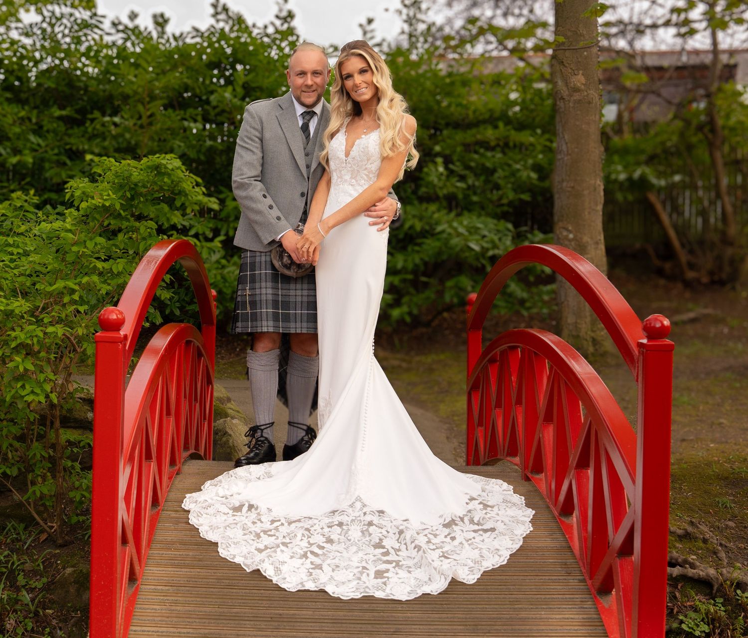 A couple poses on a red Japanese garden bridge, with a white lace wedding dress train and traditional Scottish kilt attire.