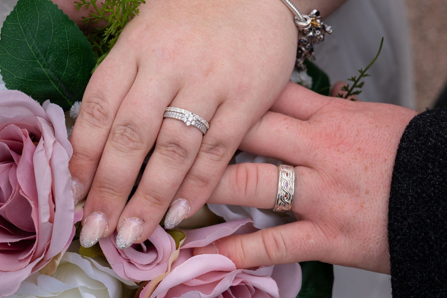 Two hands with wedding rings rest together against pink roses, showcasing elegant silver bands and a diamond engagement ring.
