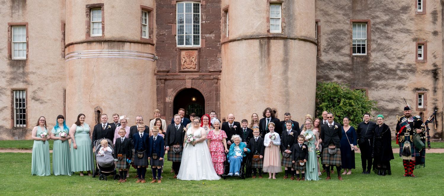 Large wedding party gathered in pastel bridesmaid dresses and formal attire outside a stone castle for group photo.