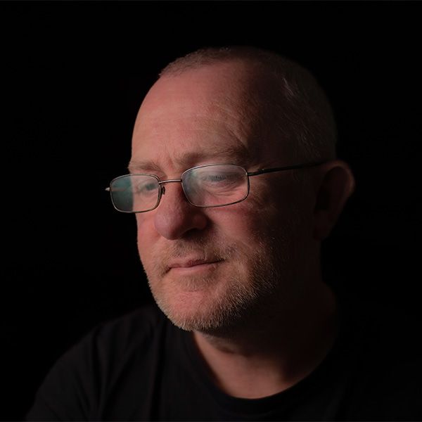 Dark portrait photo of a contemplative figure wearing glasses and a black shirt against a black background.