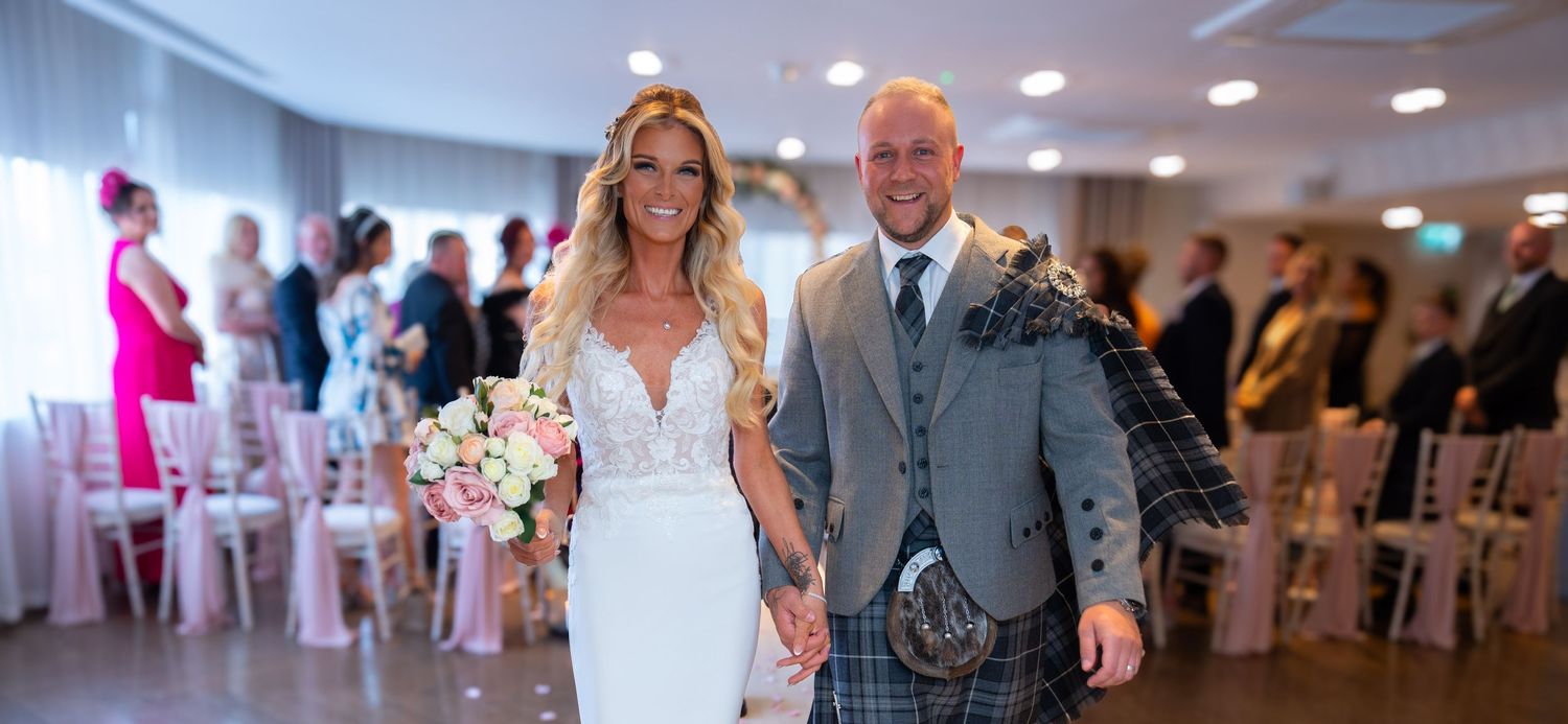 A couple walks down the aisle at their wedding reception in a bright indoor venue with pink floral decorations.