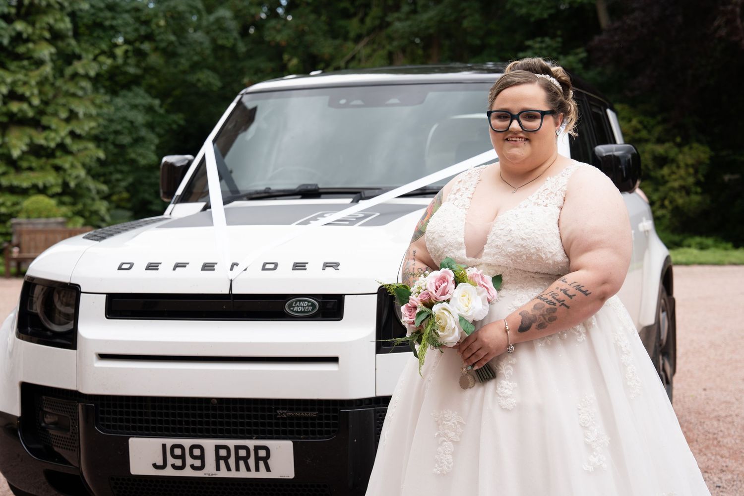 A bride in a white lace dress holding a pink bouquet poses in front of a white Land Rover Defender vehicle outdoors.