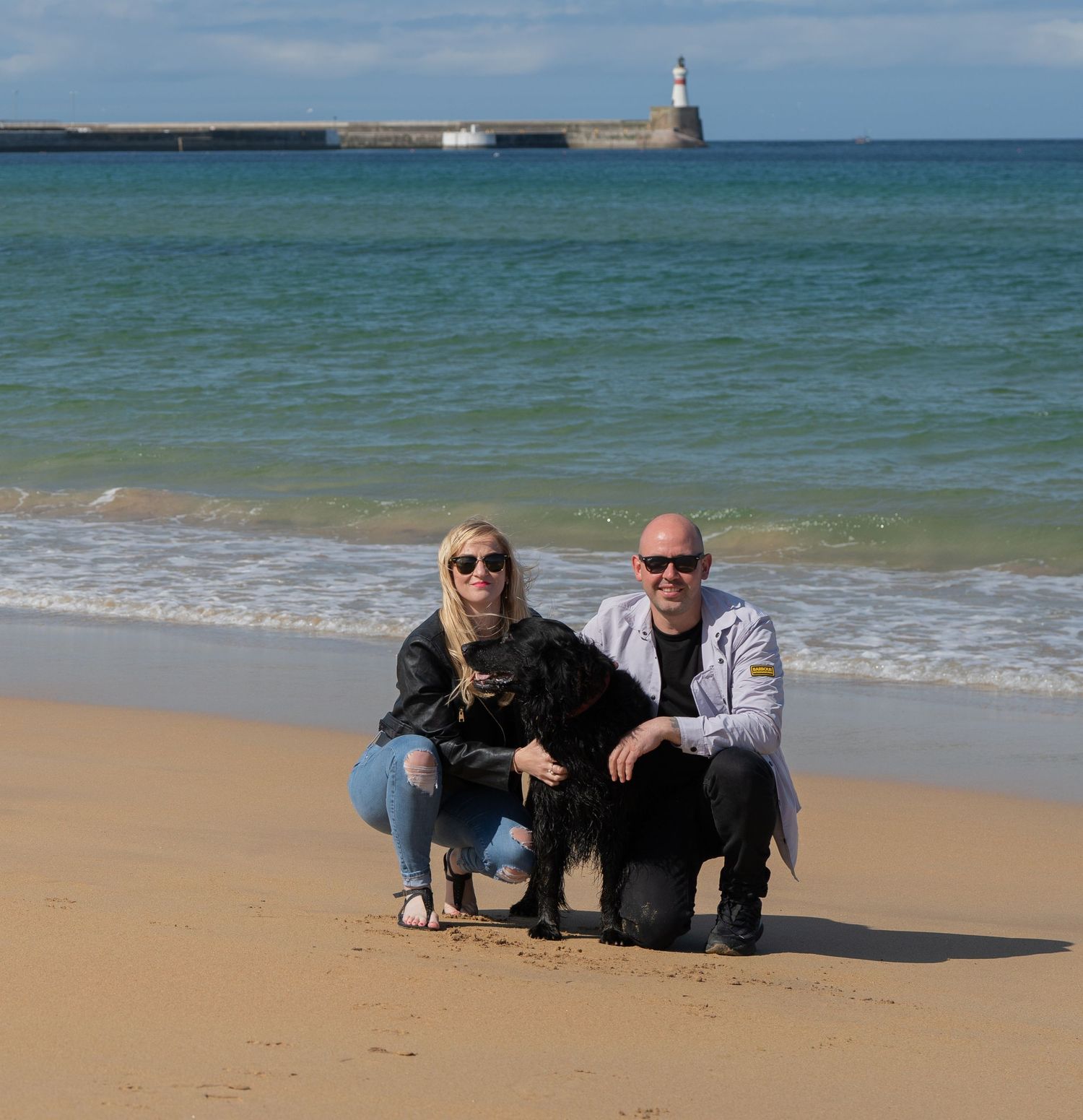 Two people kneeling on sandy beach with small black dog and lighthouse visible in turquoise waters behind them.