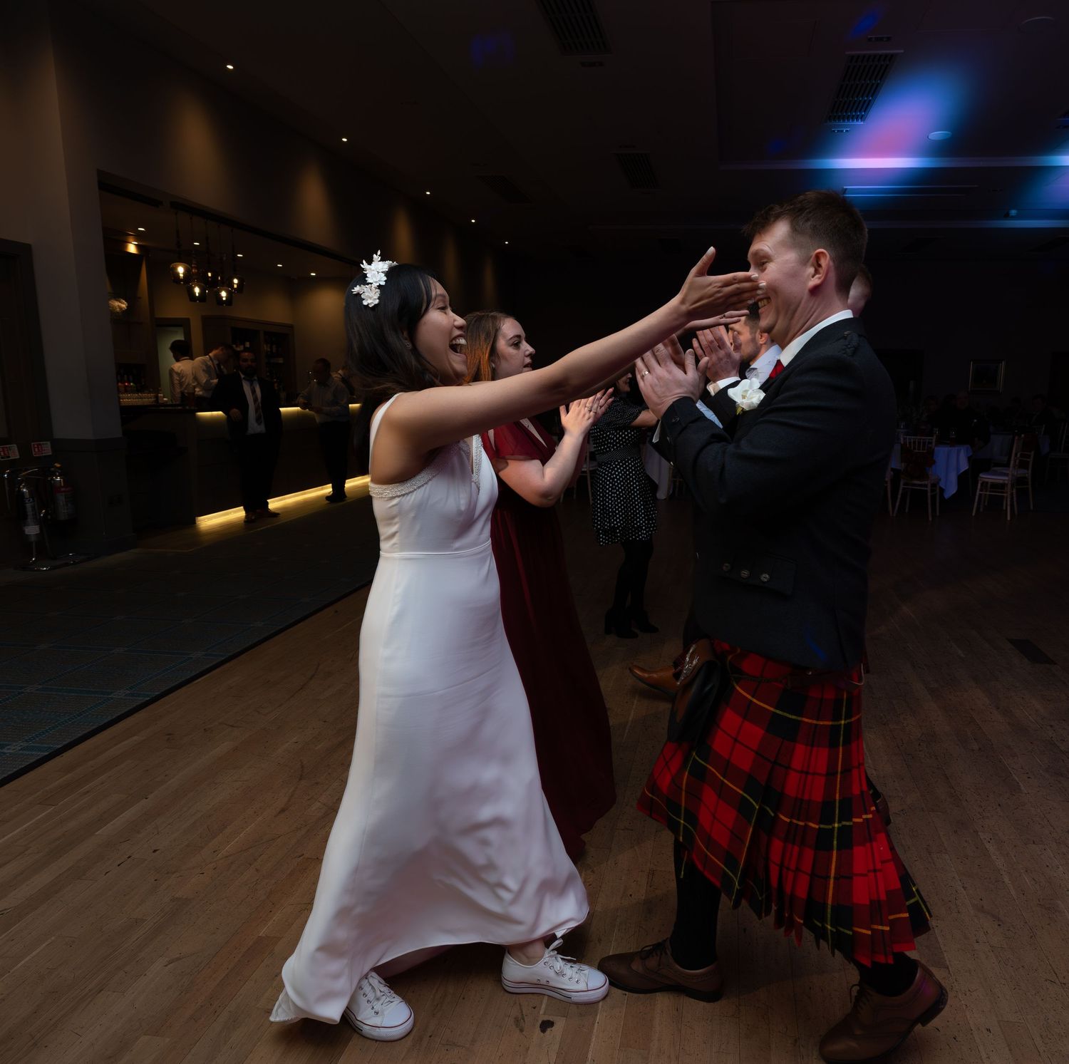 A couple dances energetically at their wedding reception, with the groom wearing a traditional red tartan kilt.
