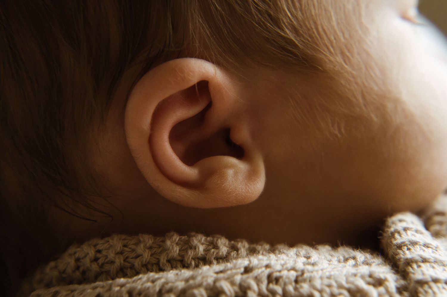 Close-up view of a baby's ear and blonde hair against a cream knit sweater in warm, natural lighting.