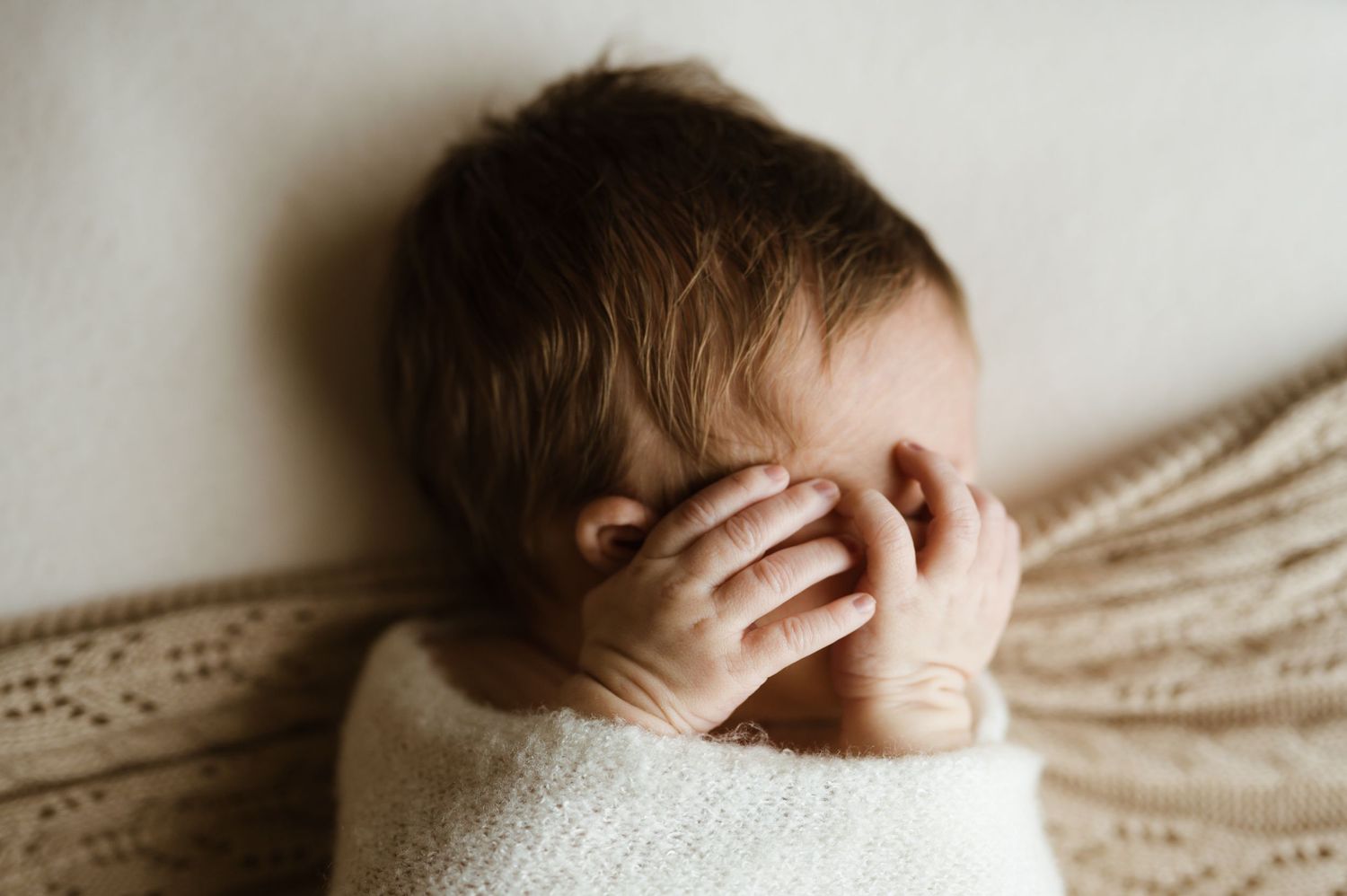 A swaddled infant lies on beige fabric while covering their eyes with their hand in a candid moment.