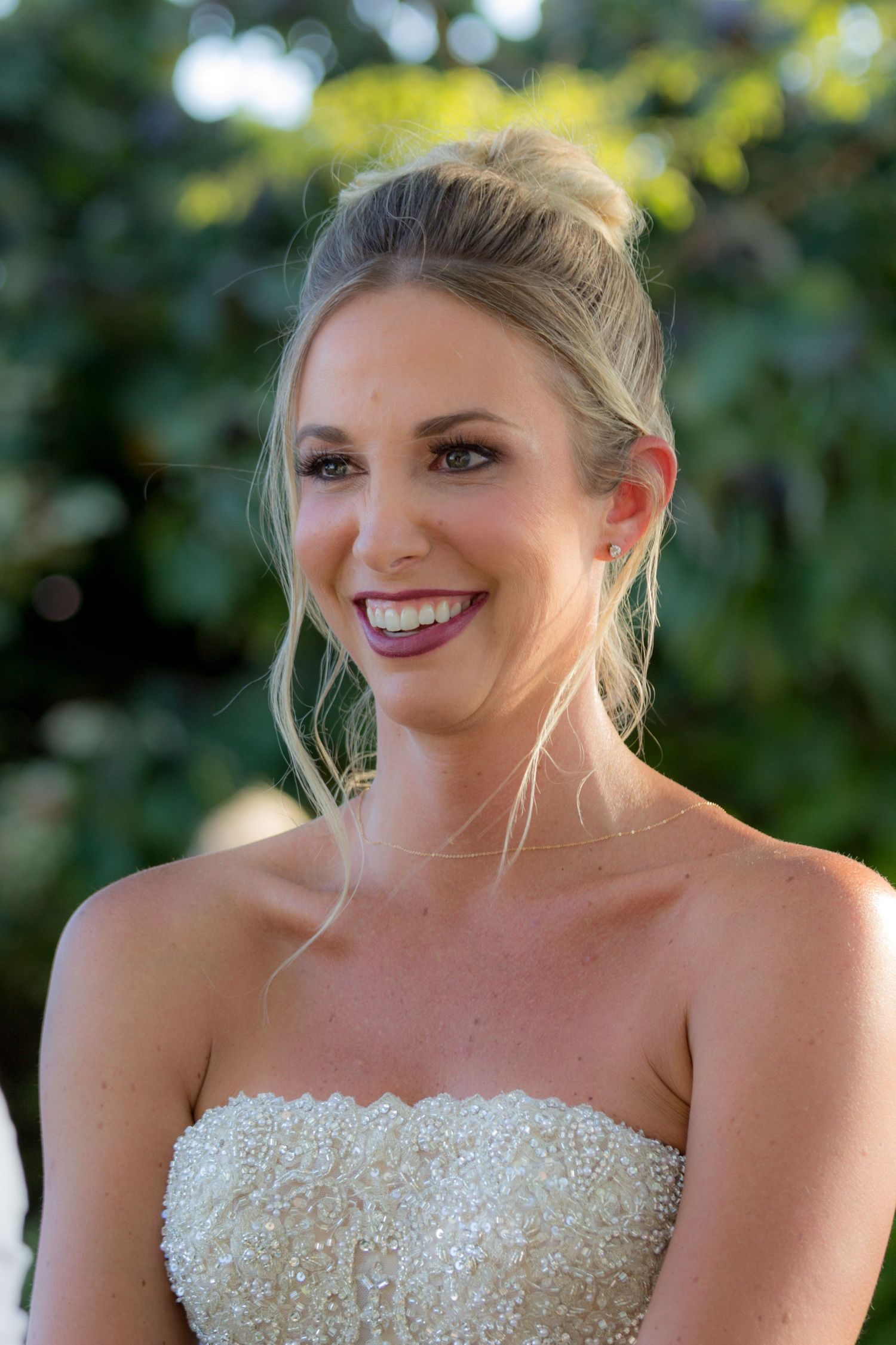 A bride in a sequined strapless wedding dress smiles outdoors with a blurred natural background.