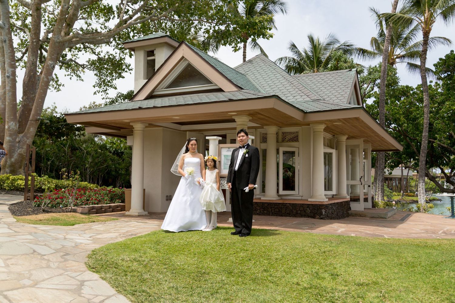 A wedding couple poses outside Hilton Waikoloa Village wedding chapel surrounded by palm trees.