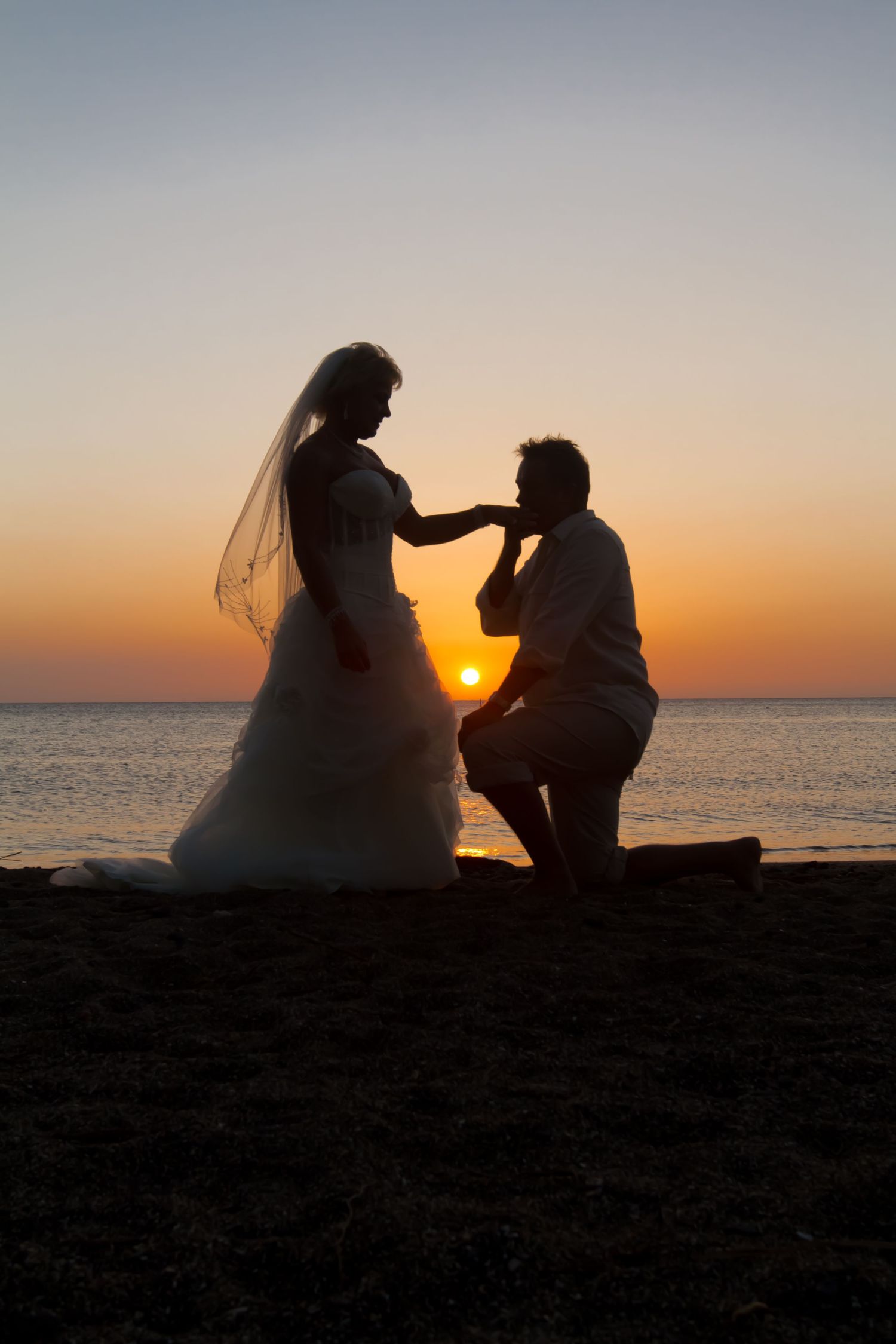 A silhouetted proposal scene at sunset with ocean backdrop as he kneels before bride in wedding dress at Mauna Lani Bay near Kailua Kona.