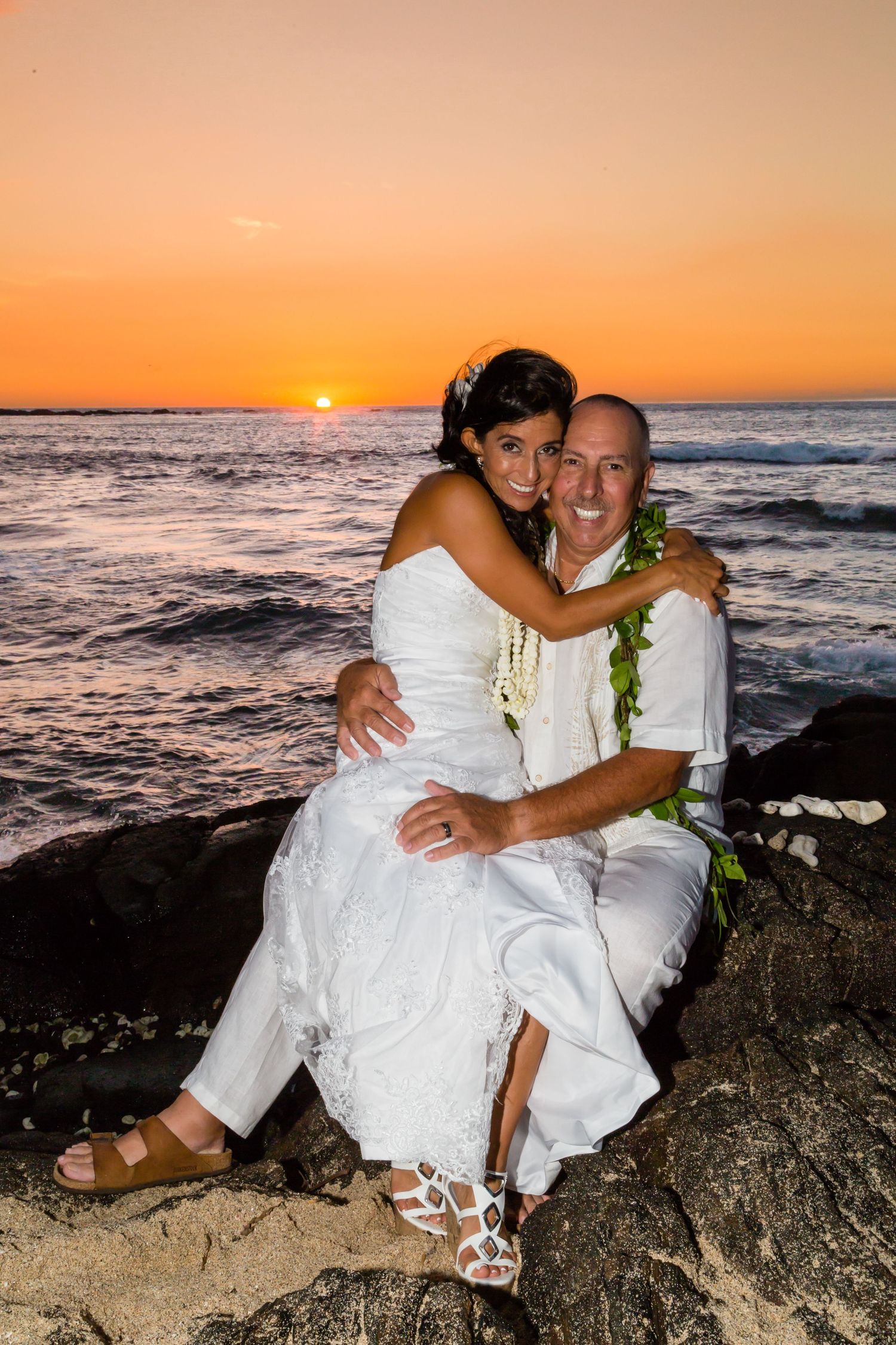 A couple embraces at Kukio beach in Kailua-Kona, Hawaii, at sunset, wearing traditional Hawaiian wedding attire.