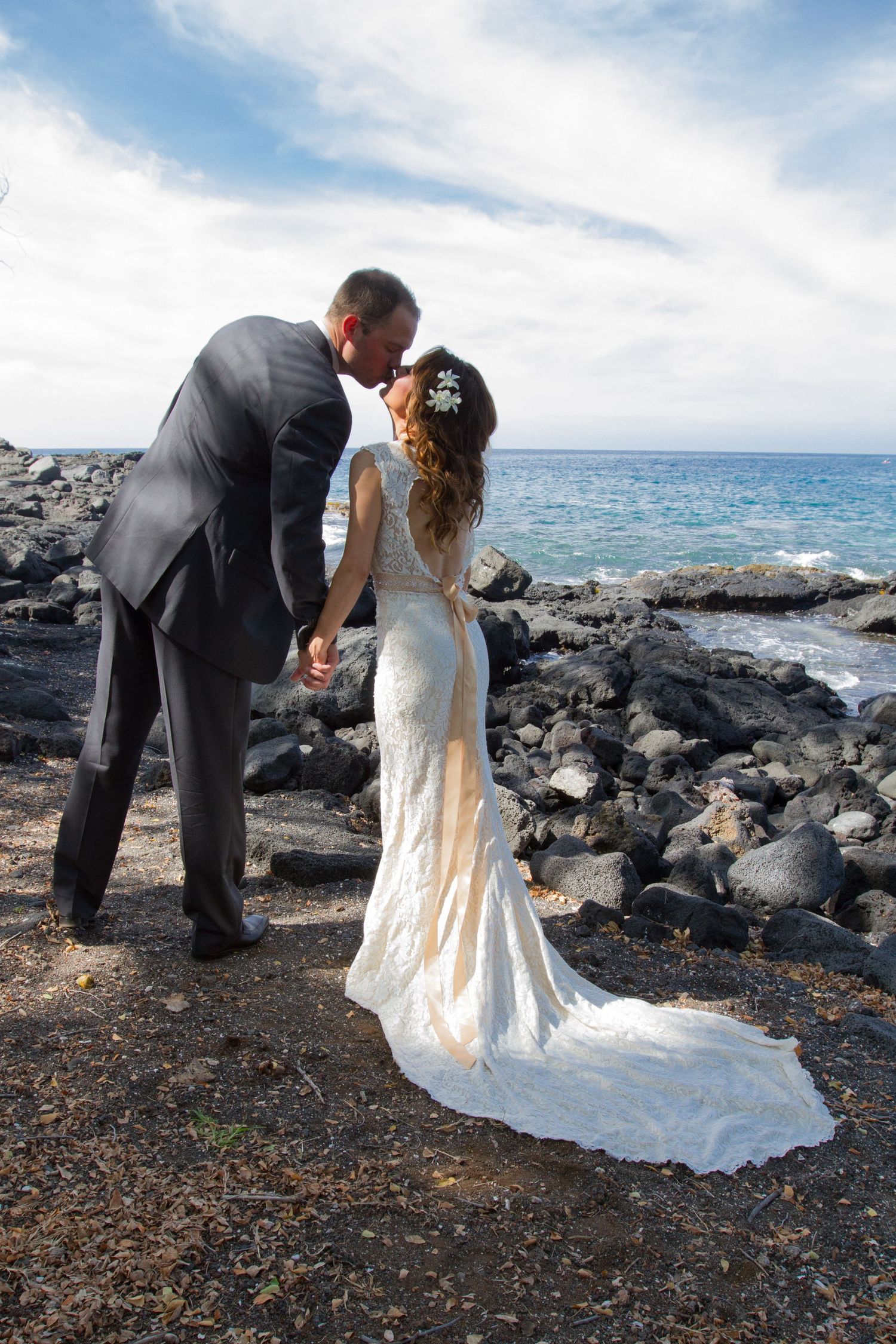 Romantic wedding couple shares a moment on rocky volcanic shoreline in Hawaii with ocean waves and blue sky backdrop.