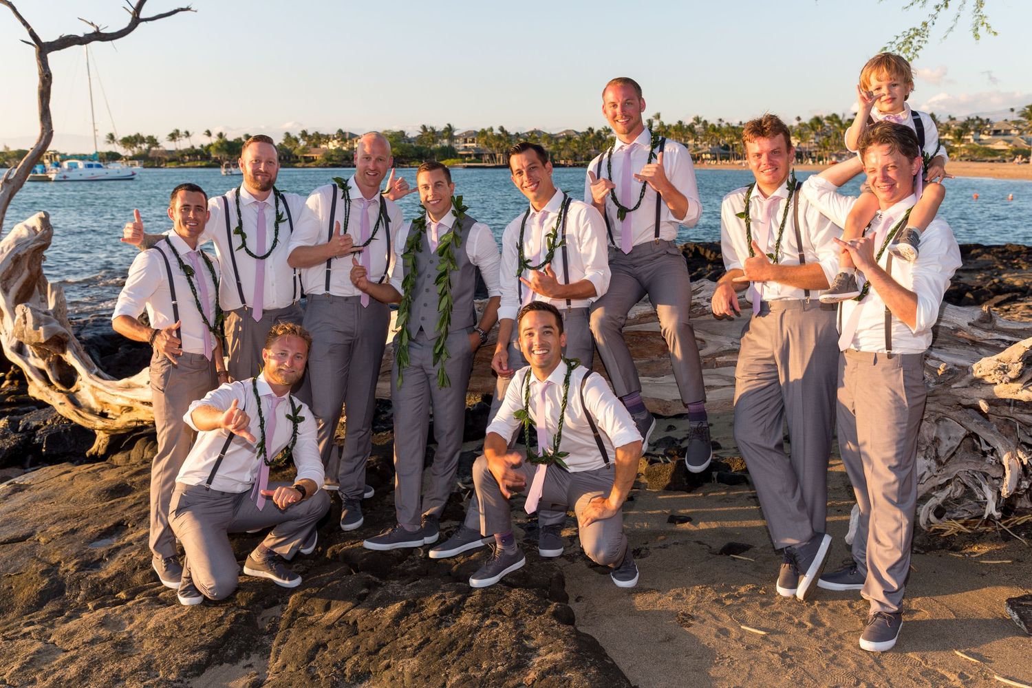 Wedding party in light gray suits and suspenders pose together on a rocky beach at sunset.