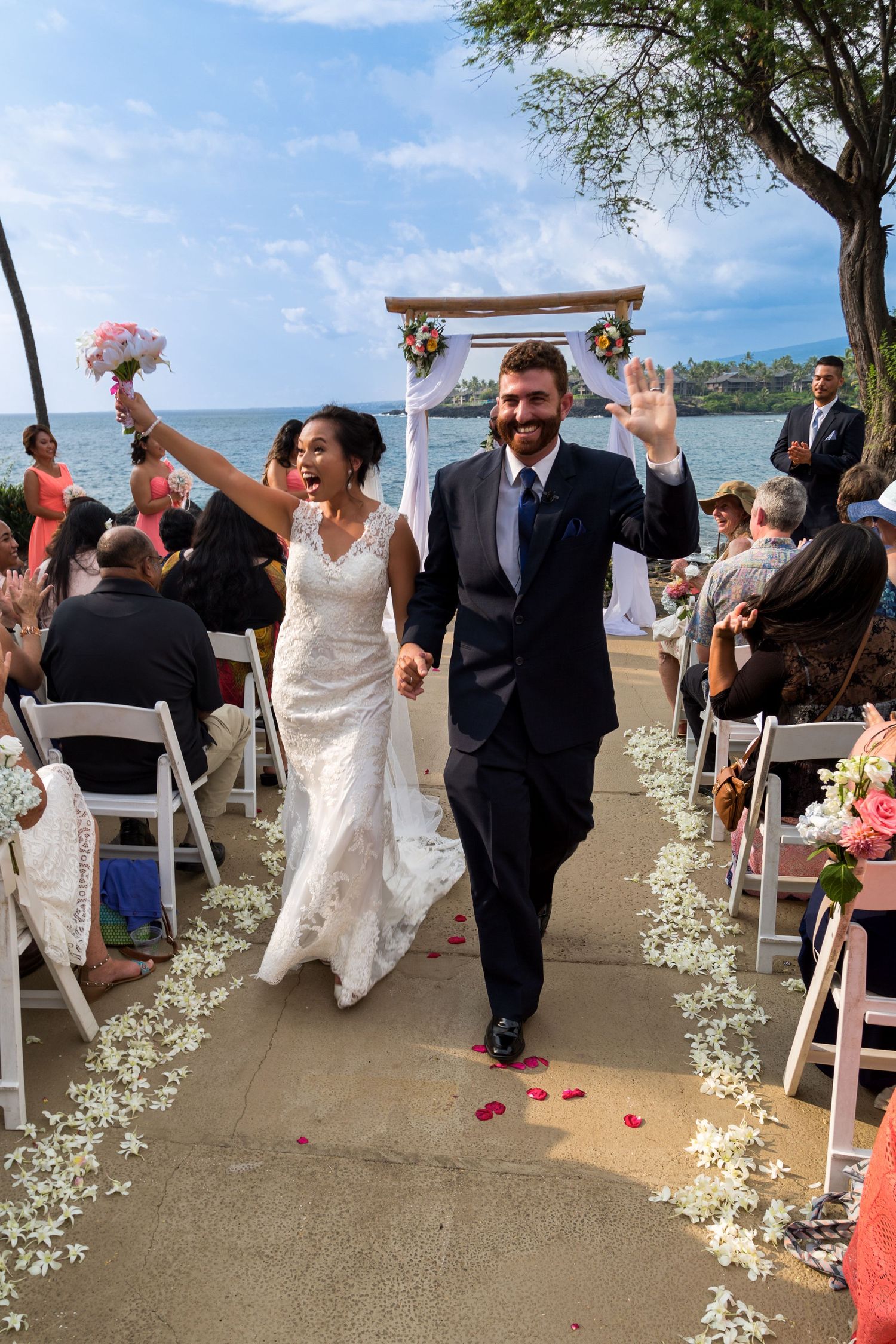 Newlyweds walk down the aisle decorated with flower petals under a blue sky by the ocean at the lawn of the Outrigger Kona Resort & Spa.