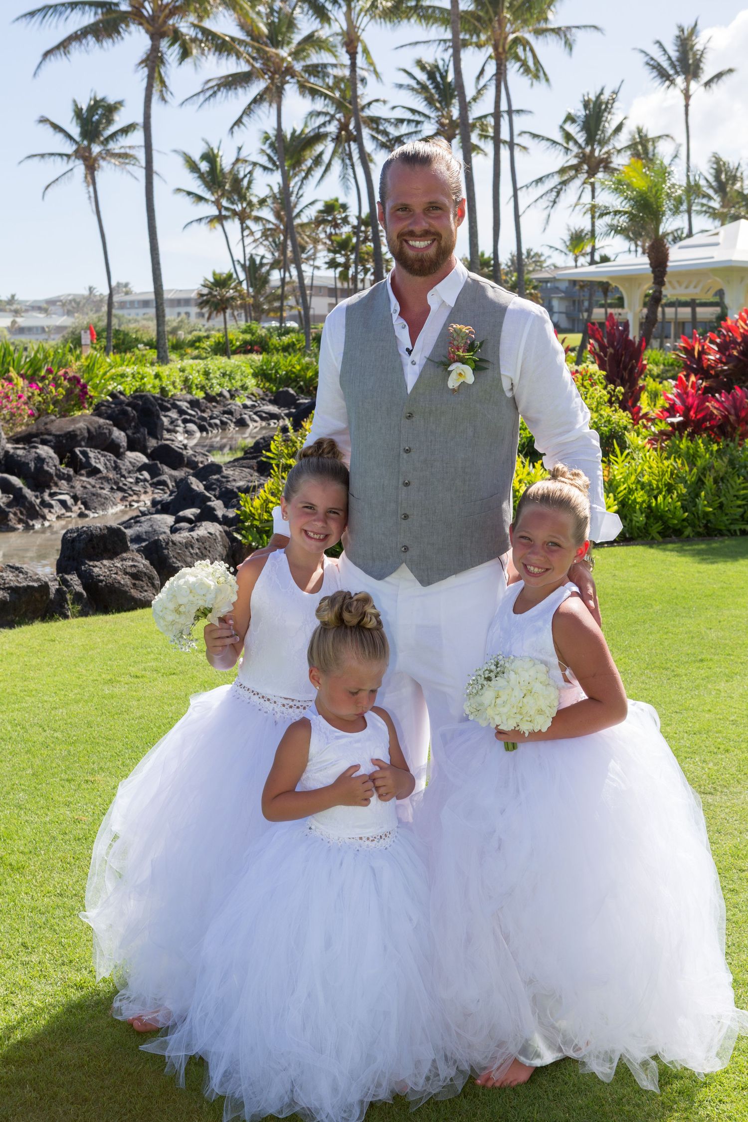 Three flower girls in white tulle dresses pose with a groomsman in a wedding at Princeville in Kauai.