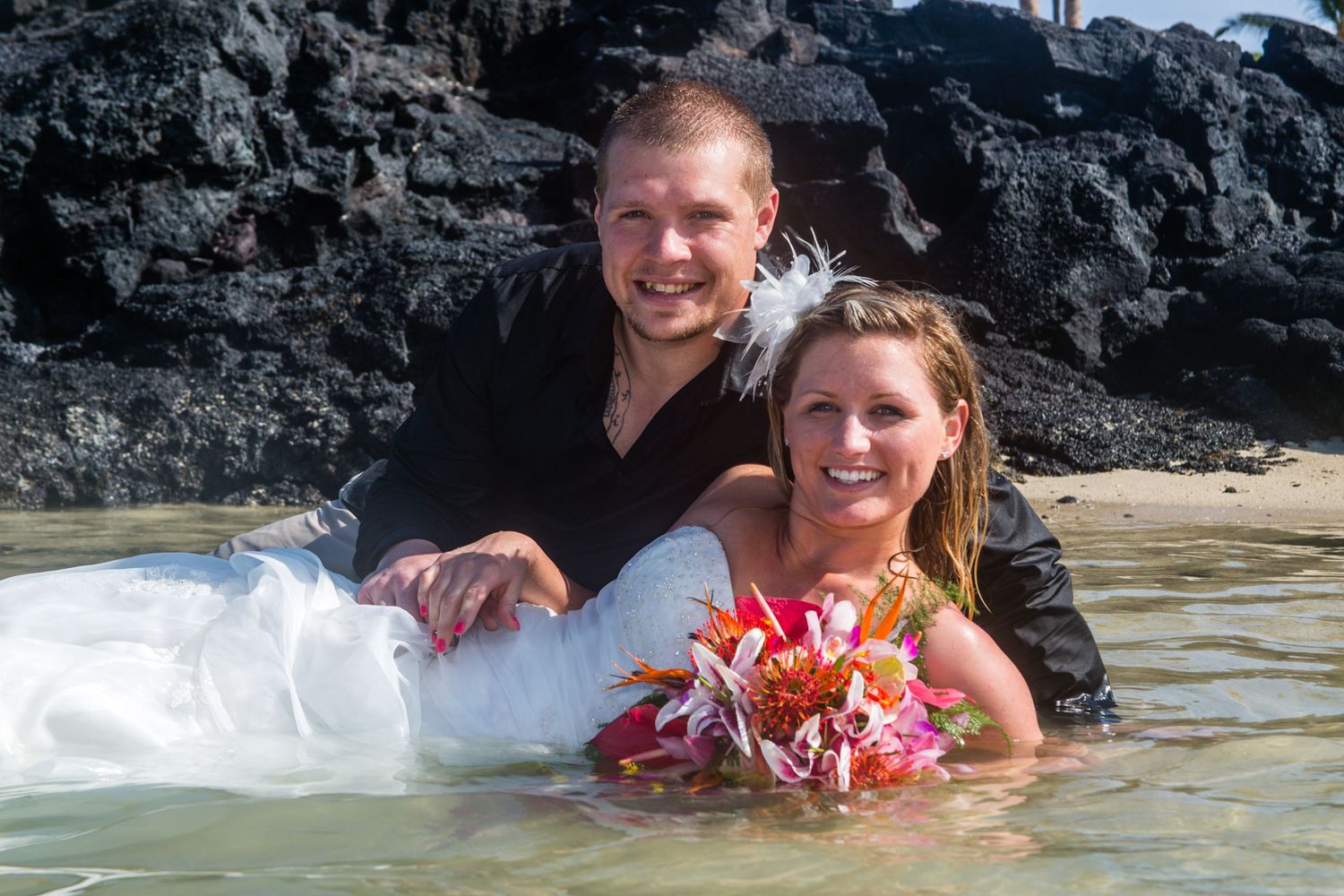 A couple in wedding attire poses together while sitting in shallow tropical water with flowers and rocky cliffs behind them at Kikaua beach.