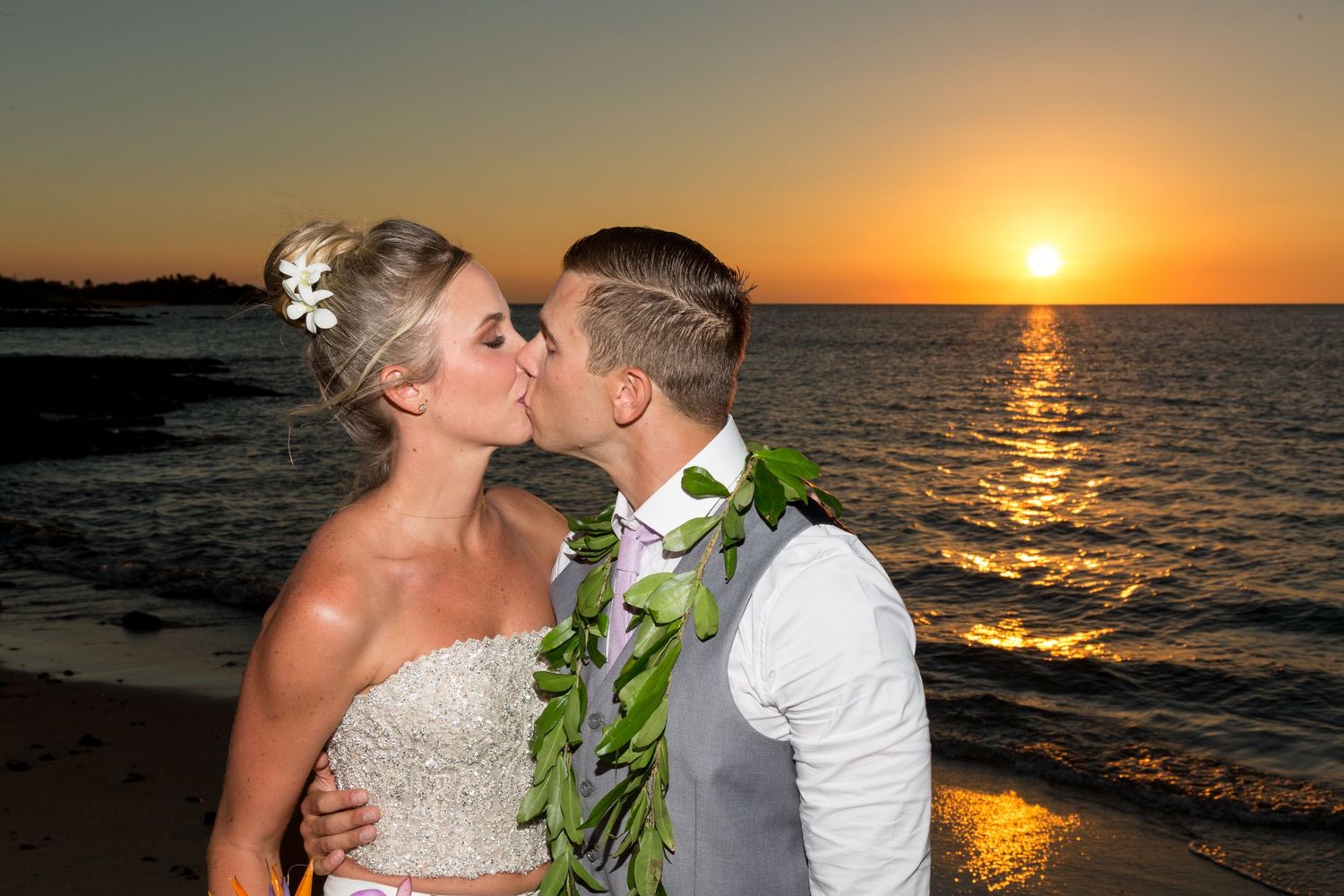 A couple shares a romantic sunset kiss on a beach with a traditional Hawaiian lei around the groom's neck.