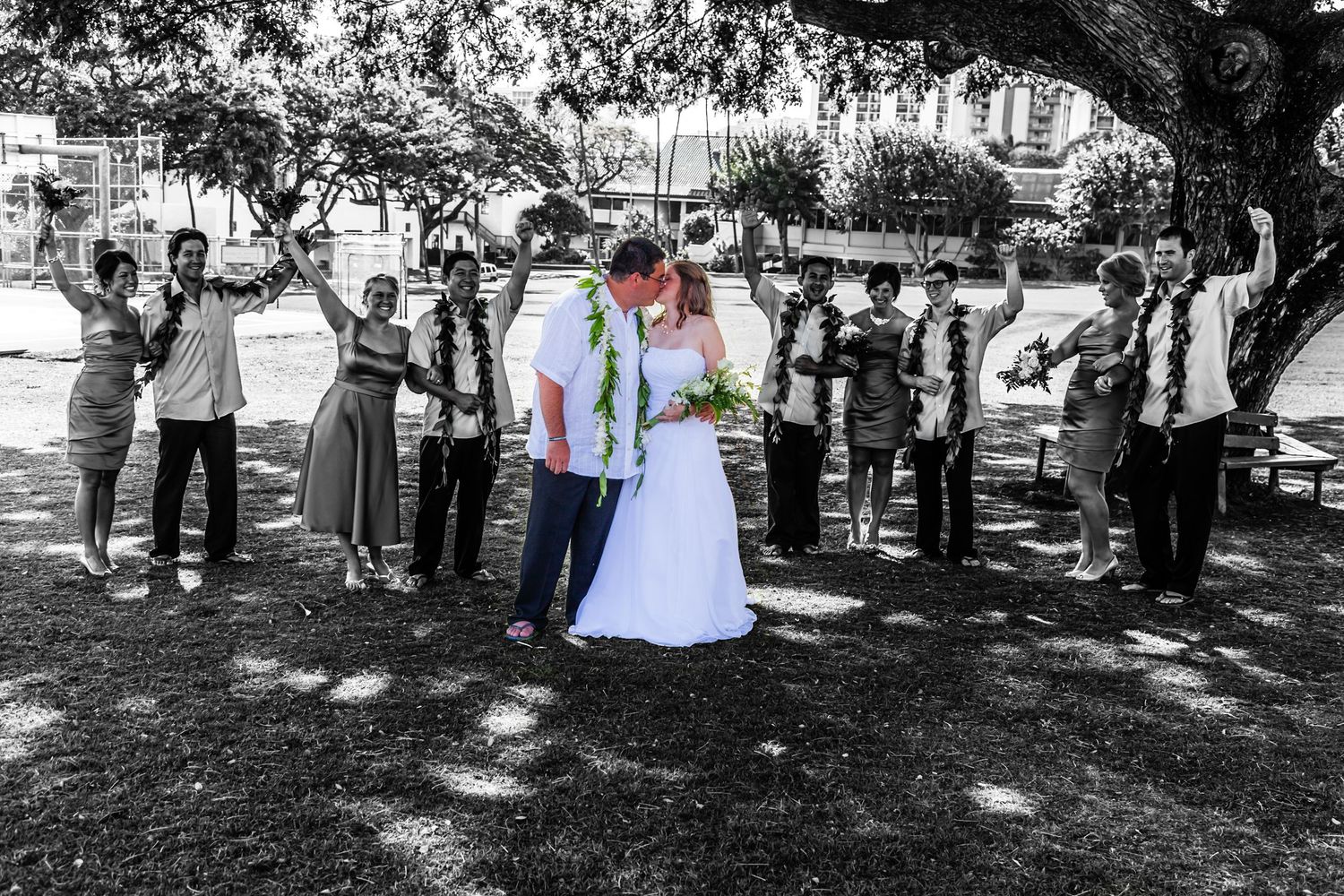 A black and white wedding photo showing a couple surrounded by guests under the shade of a large tree in a park in Honolulu, Hawaii.