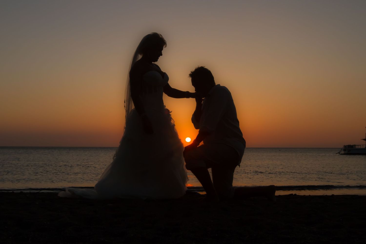 Silhouetted figures sit on beach at sunset with vibrant orange sky reflecting off calm ocean waters.