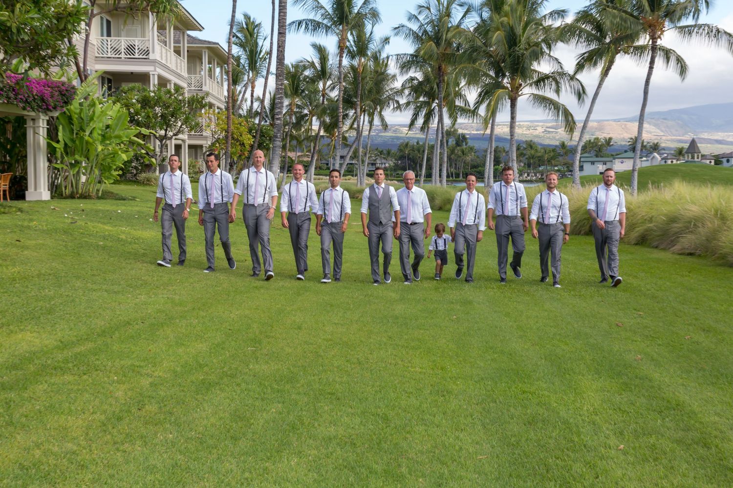 Wedding party wearing blue and white stands in a line on lush green grass with palm trees and ocean view in Hawaii.