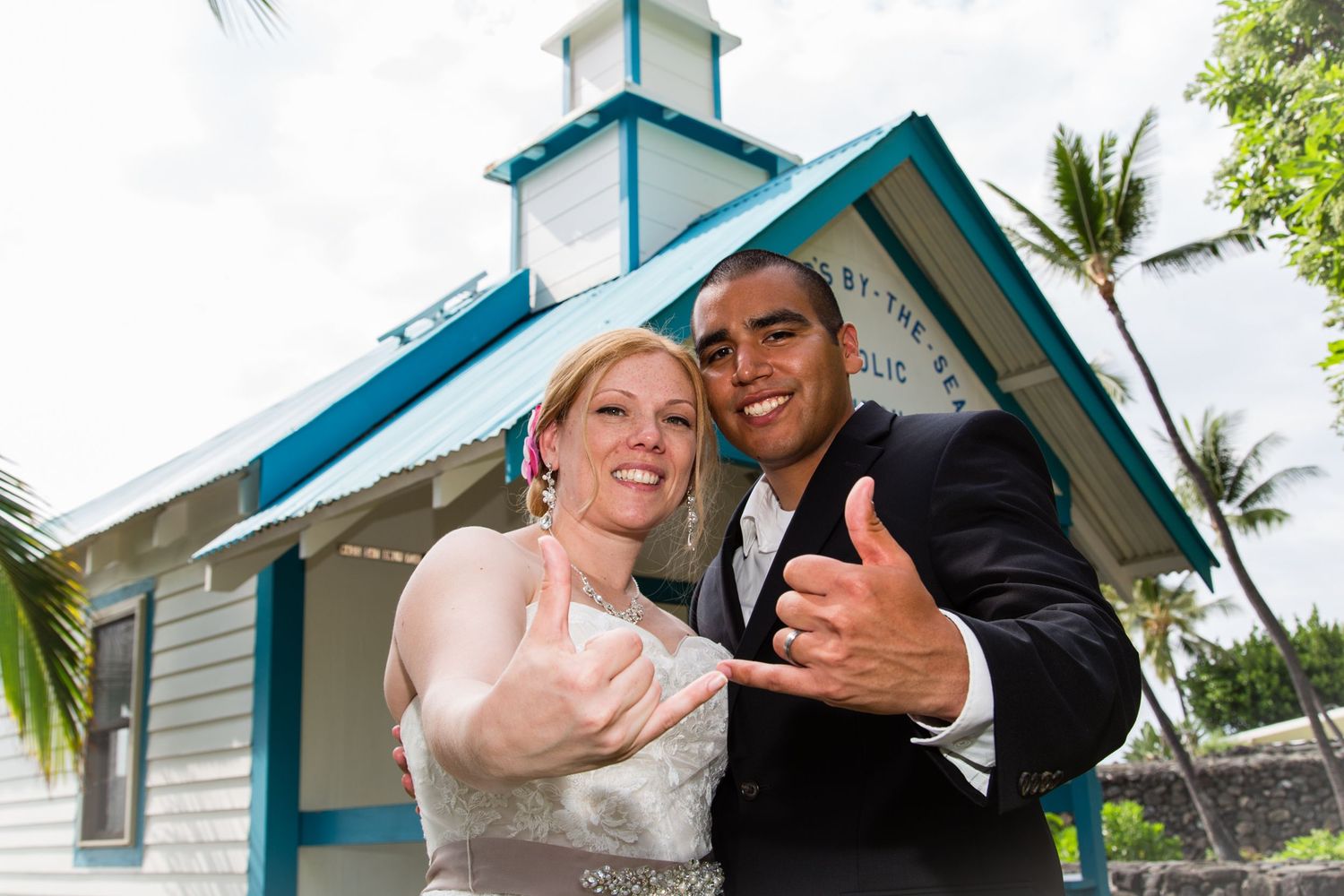 A joyful wedding couple gives the shaka sign in front of the blue and white Catholic Saint Peter's chapel in Kona, Hawaii.