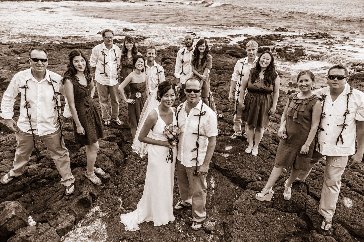 Sepia photo of wedding party posing on volcanic rocks in casual beach attire next to the Living Stones Church in Kailua Kona, Hawaii.