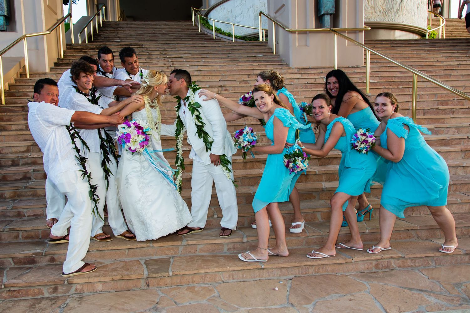 At the Hilton Waikoloa Village hotel in the Big Island. a playful wedding party poses for a tug-of-war style photo on outdoor steps.
