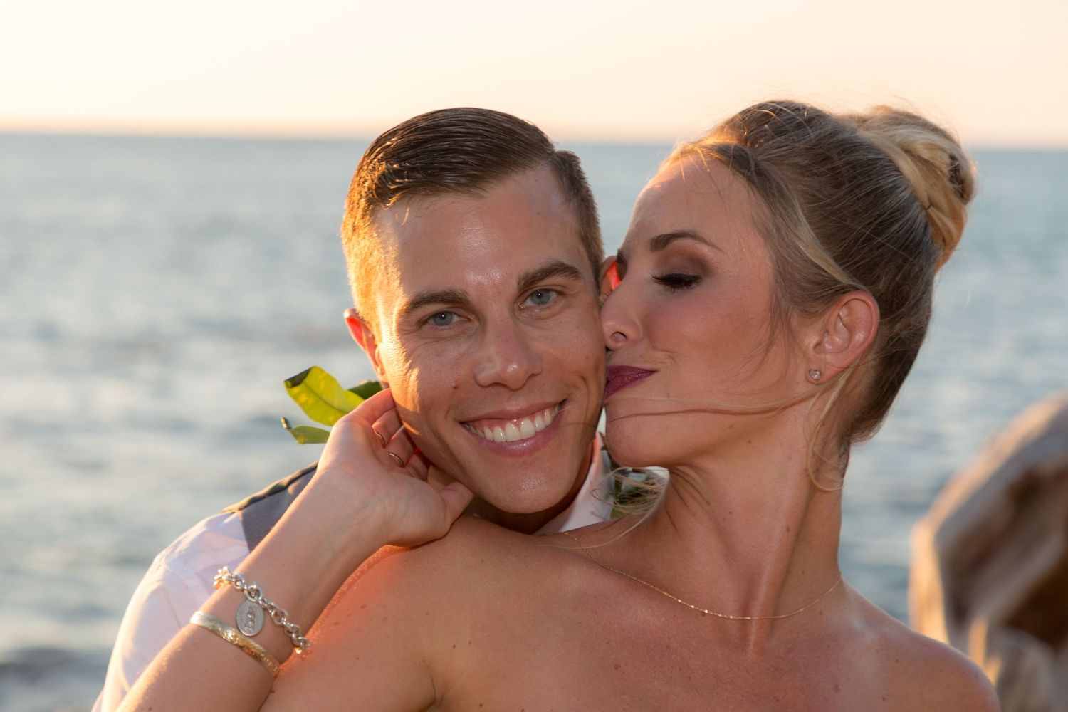 A romantic couple embraces at sunset on a beach with an orange flower accent in their hair.