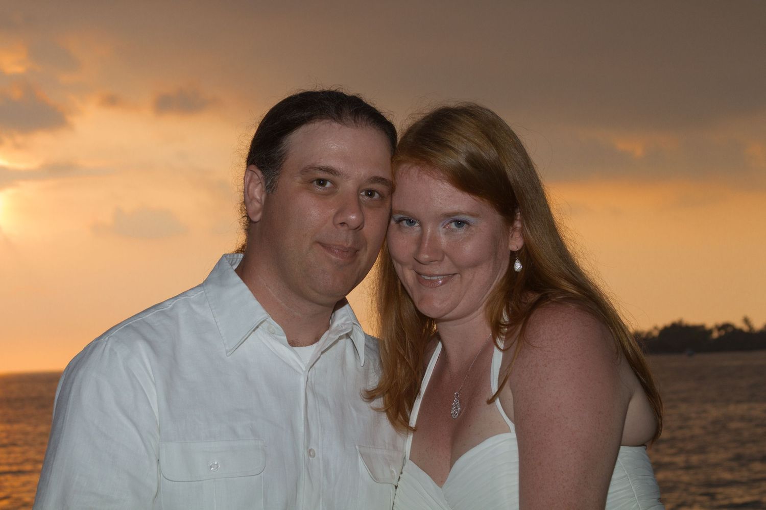 A wedding couple pose together at sunset by the ocean while wearing white attire.