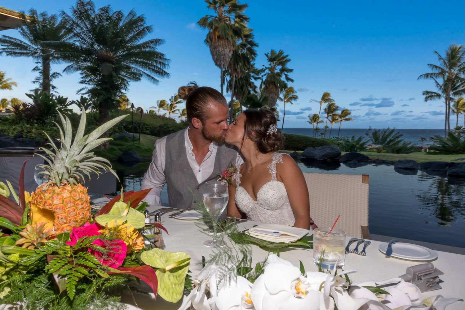 Newlyweds share an intimate moment at a reception table decorated with colorful flowers and palm trees during a wedding in Kauai, Hawaii.