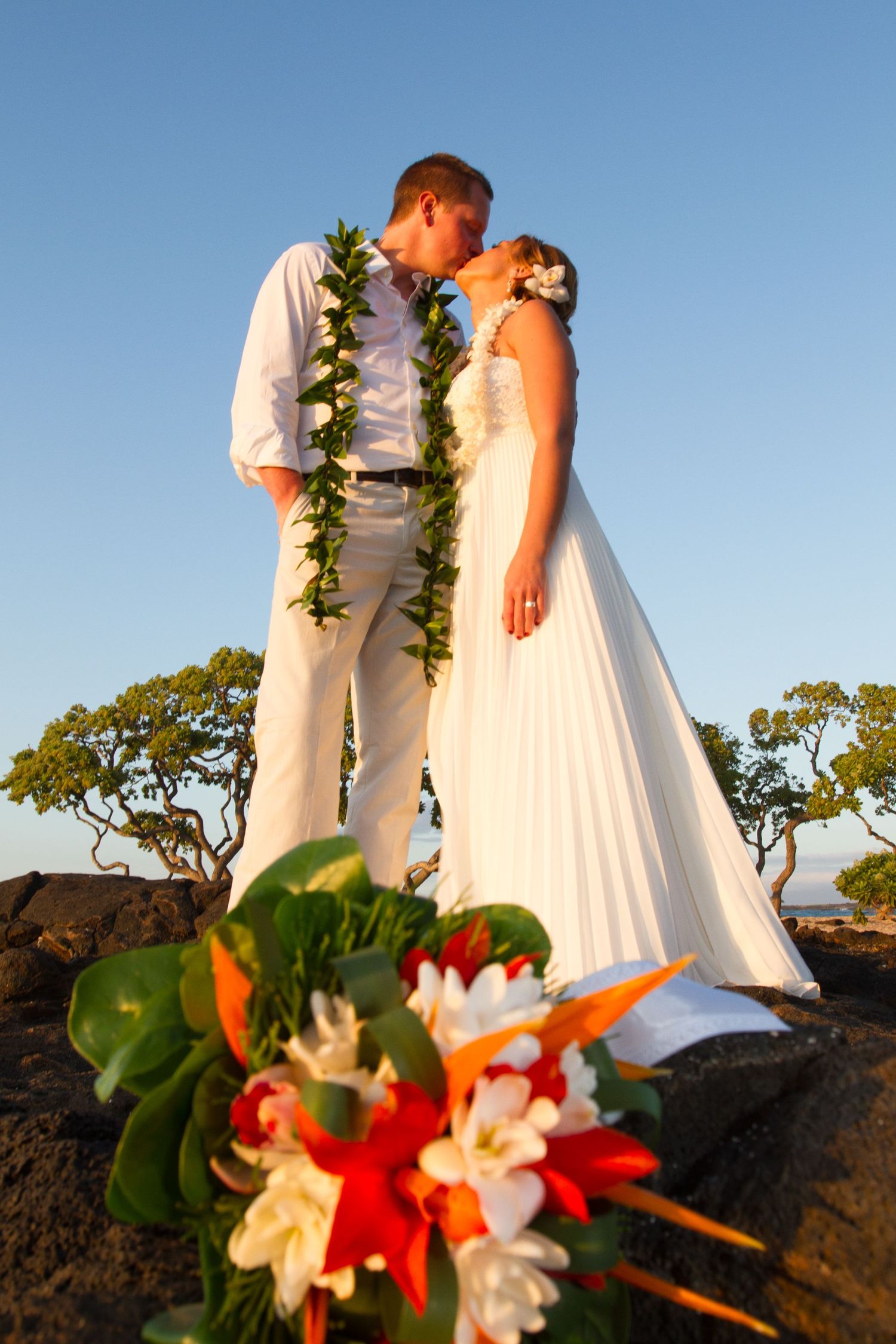 A wedding couple with leis share an intimate moment against a blue Hawaiian sky and tropical landscape at Kailua Kona, Hawaii.