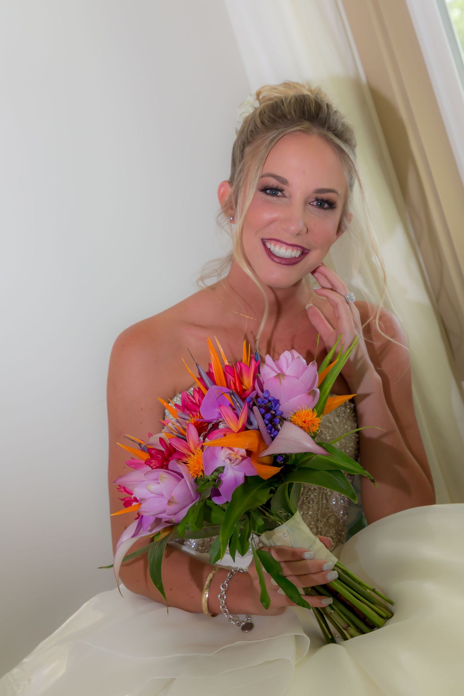 A joyful bride in a strapless white gown holds a vibrant tropical bouquet with pink, orange and purple flowers.