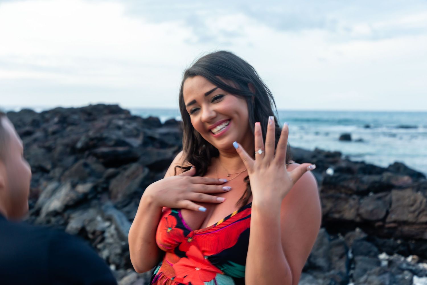 Woman in red dress shows engagement ring to camera at the Old Airport Beach Park in Kailua Kona, Hawaii after a surprise proposal.