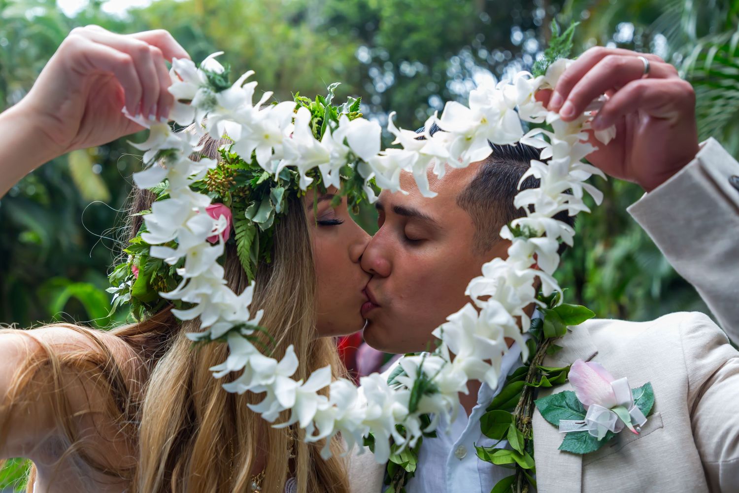 A couple shares a romantic kiss through a white flower lei forming a heart shape in a lush tropical garden setting in Kailua Kona, Hawaii.