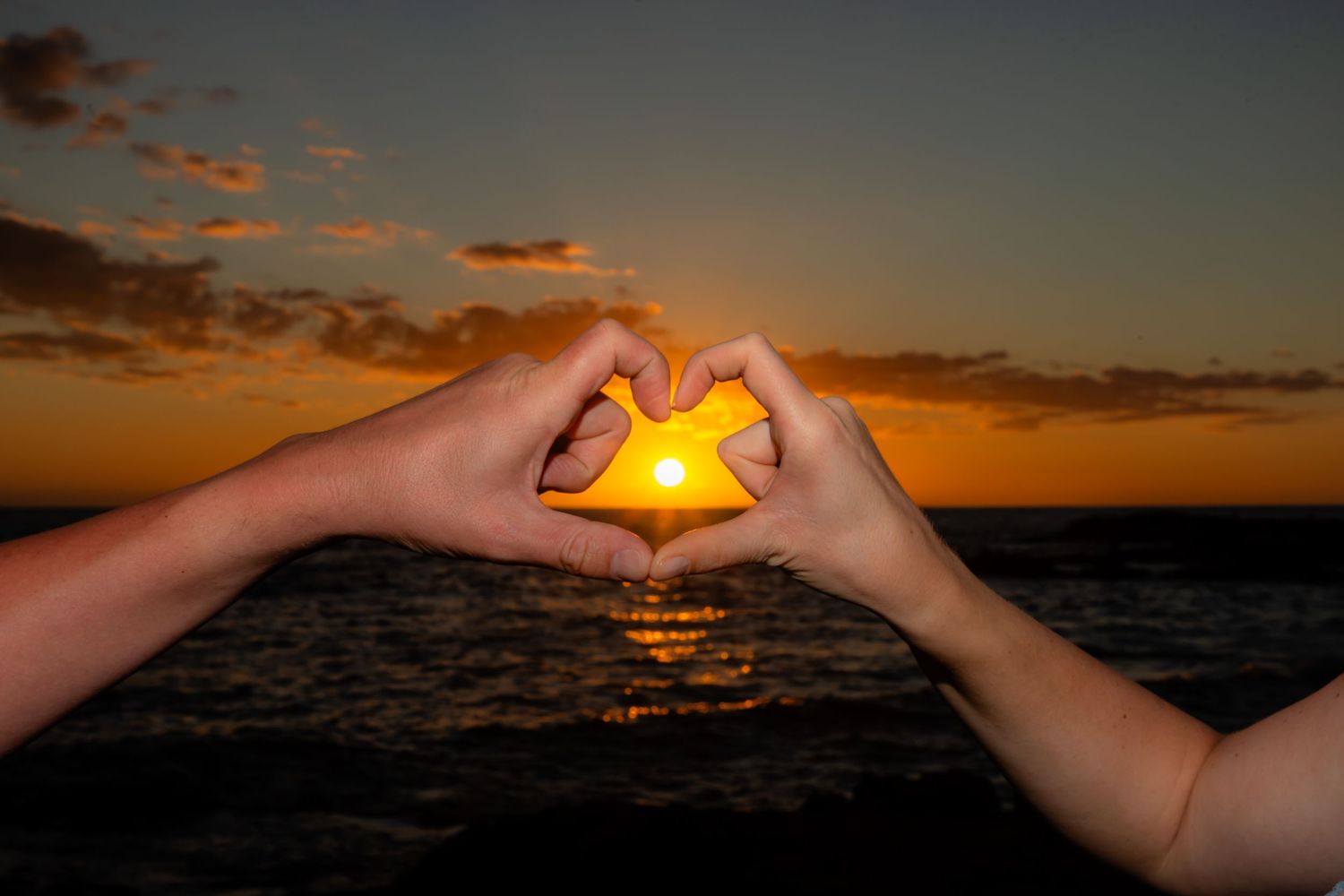 Two hands form a heart shape silhouetted against an orange ocean sunset at Kukio beach in Kailua Kona, HAwaii