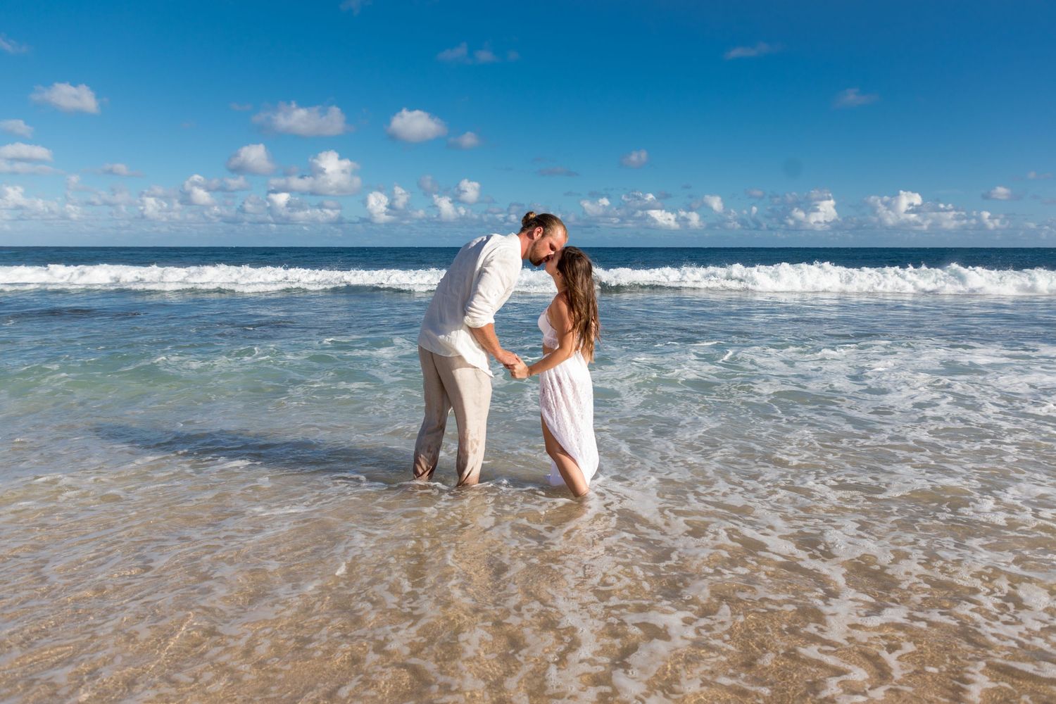 A romantic couple in white outfits embraces at the edge of the ocean waves on a sunny beach in Kauai.