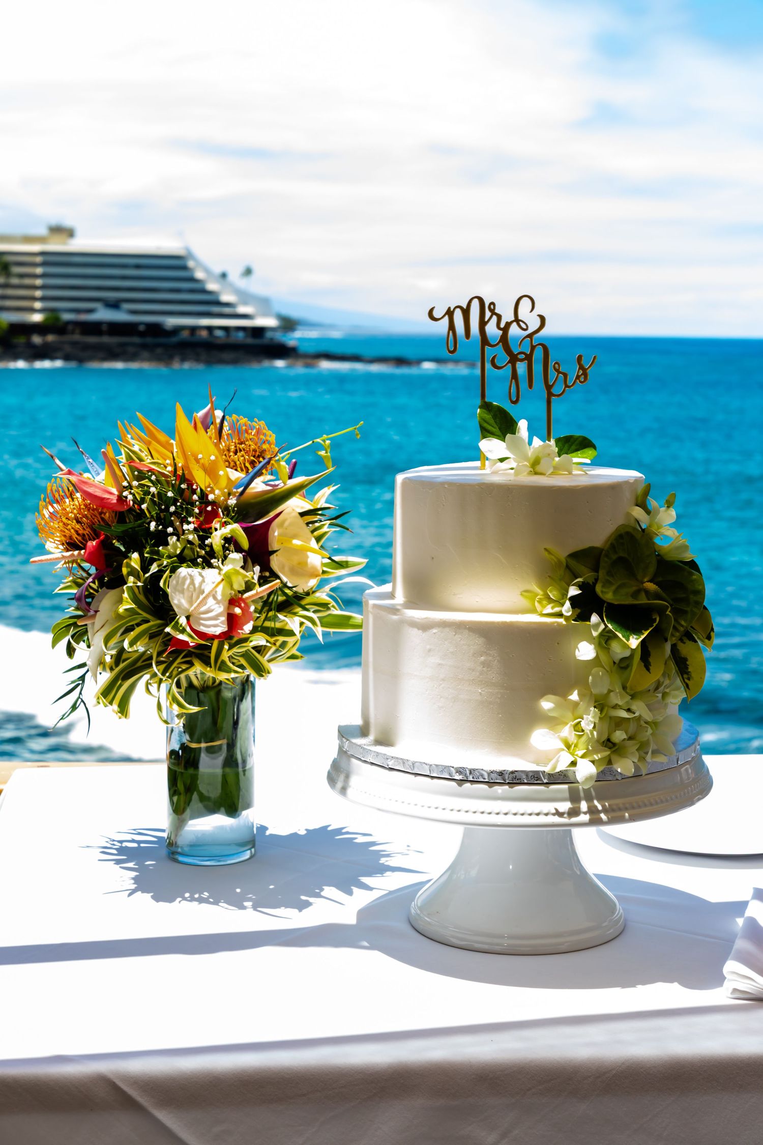 Wedding cake and tropical flower bouquet displayed oceanside with cruise ship in background in Papa Kona.