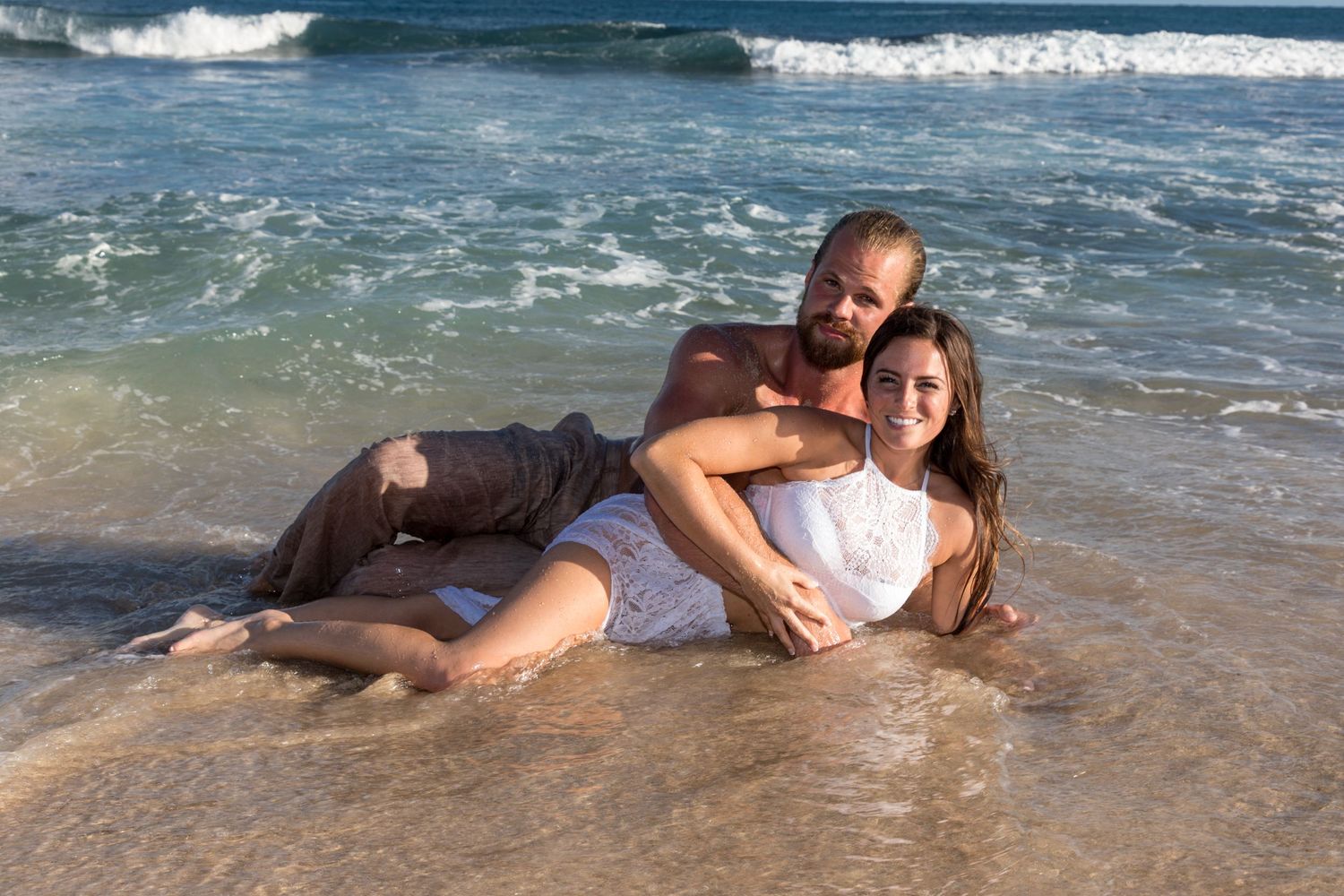 A couple lying in the surf at the beach in Kauai wearing white and blue clothing as waves crash behind them.