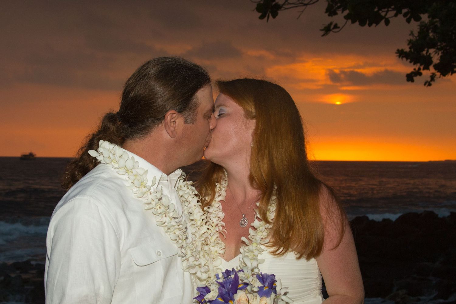 A couple shares a romantic kiss at sunset on a kona coast beach with leis and purple flowers against the orange sky.