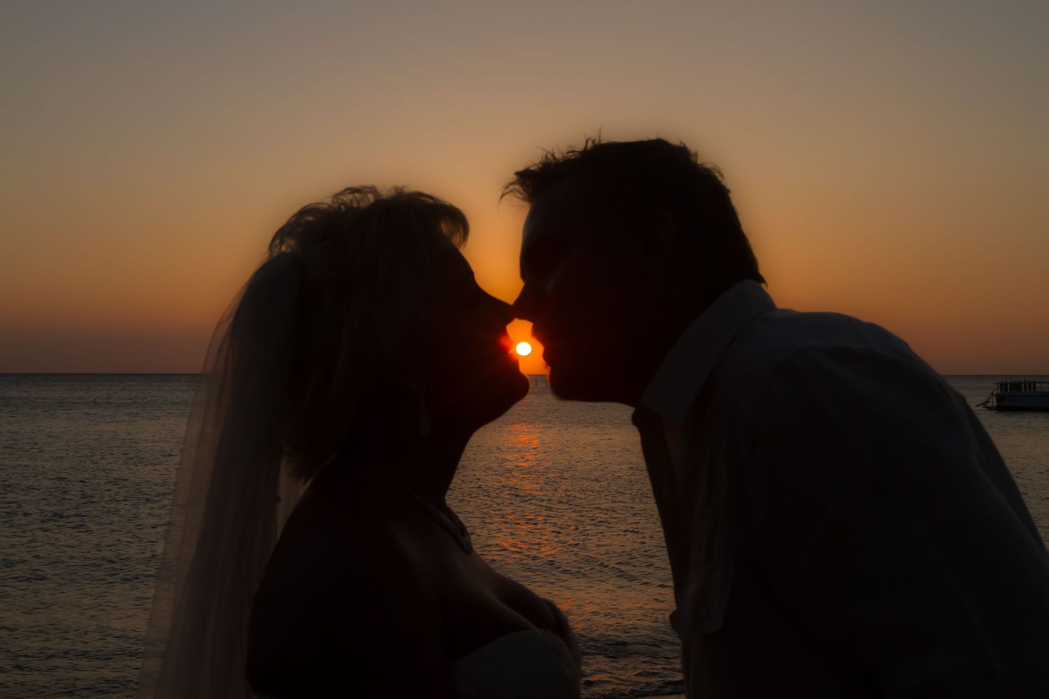 A silhouetted couple sharing a romantic kiss at sunset by the ocean with a veil blowing in the breeze.