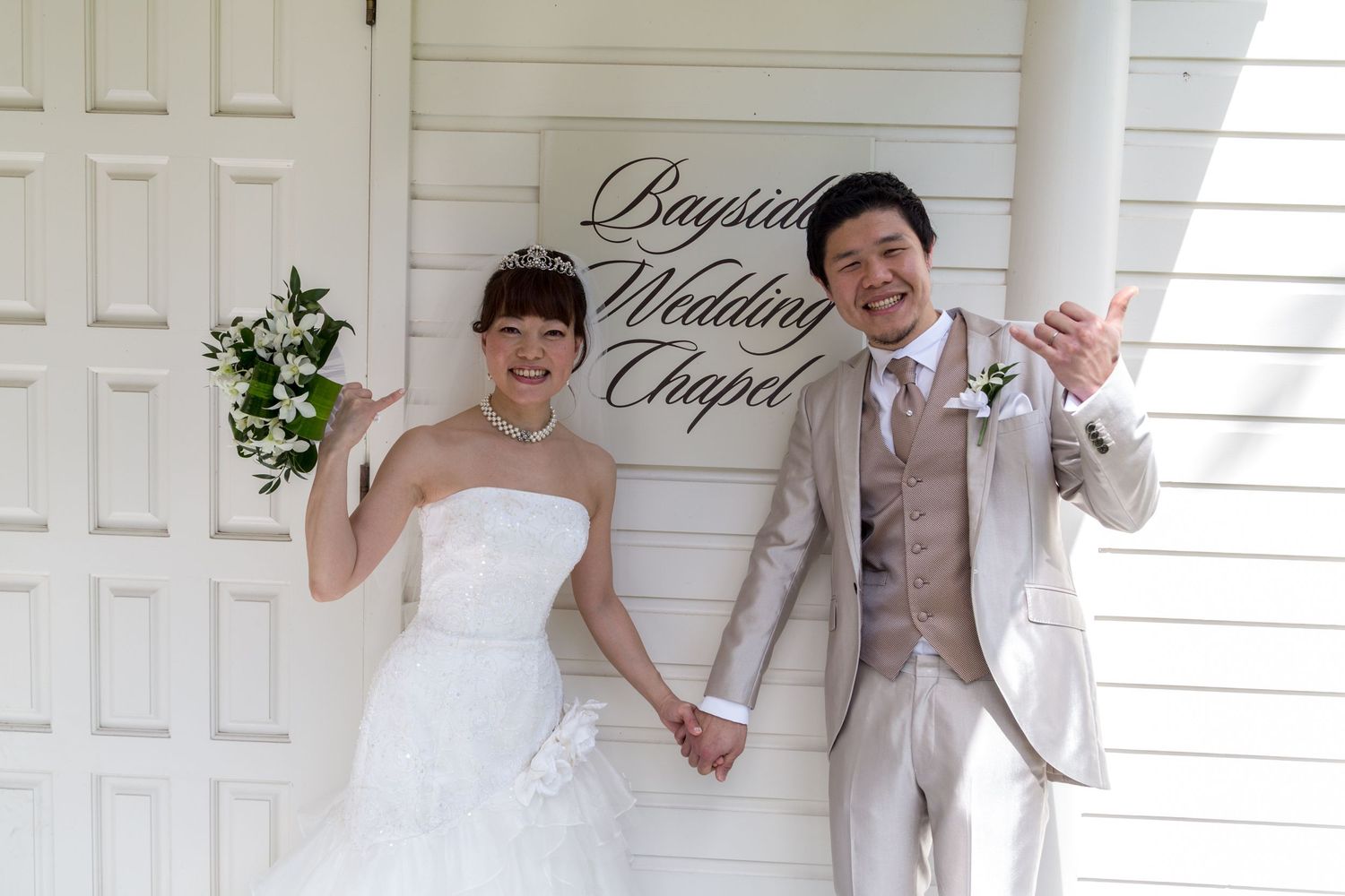 A joyful wedding couple poses outside the Bayside Wedding Chapel of the Outrigger Kona Resort & Spa, in formal attire.