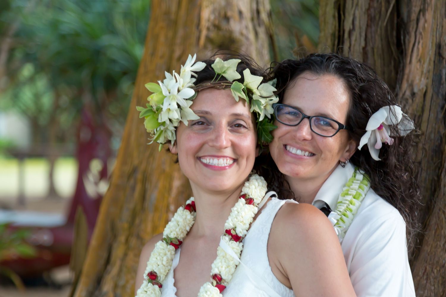 A couple wearing flower leis and white floral crowns pose against a wooden tree trunk after a same sex wedding ceremony in Kailua Kona.
