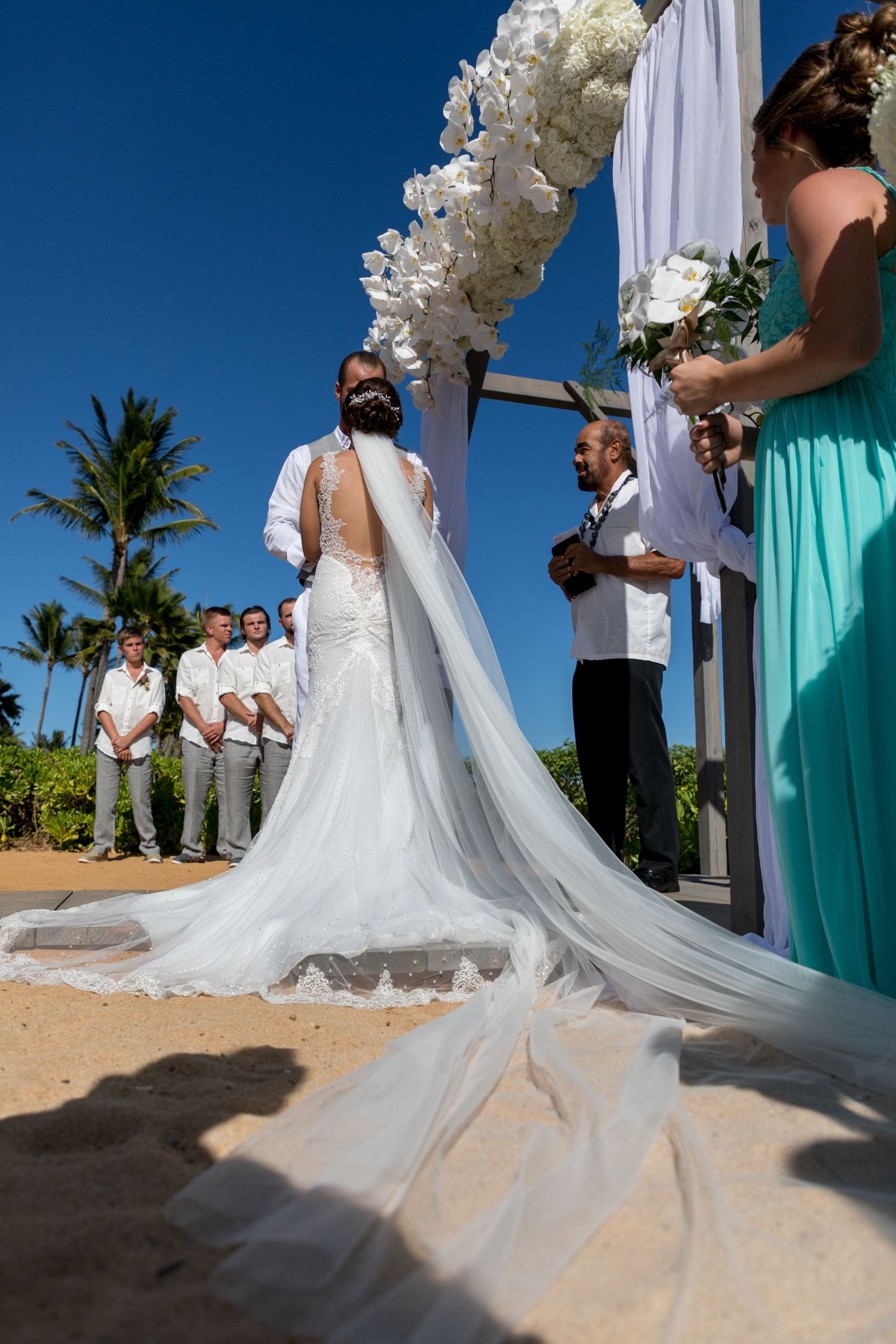 A wedding ceremony under a floral arch with flowing white dress train in foreground in Princeville, Kauai, Hawaii.