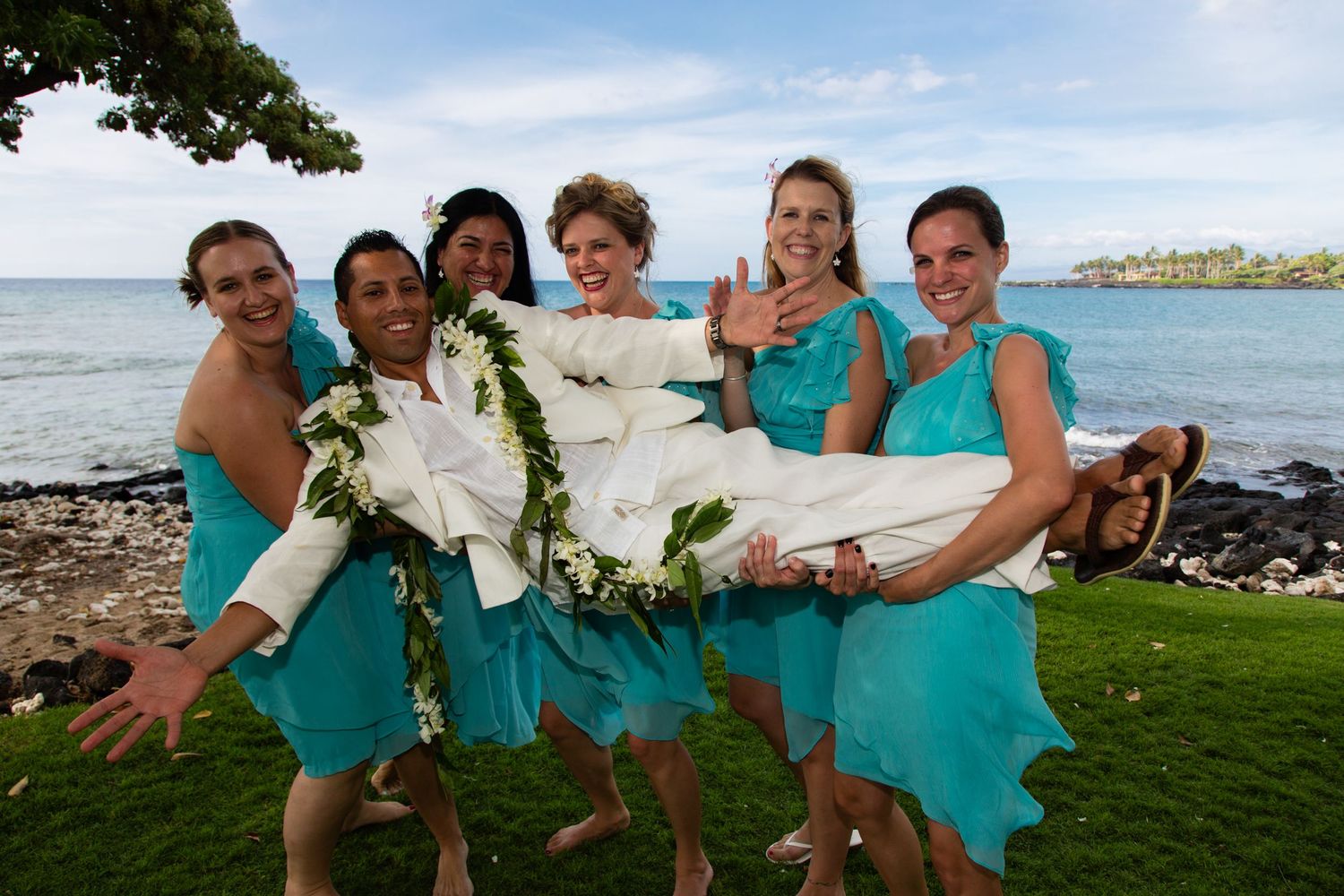Bridesmaids in turquoise dresses pose playfully by the ocean holding the groom, at Kikaua Beach Park in Kona, Hawaii.