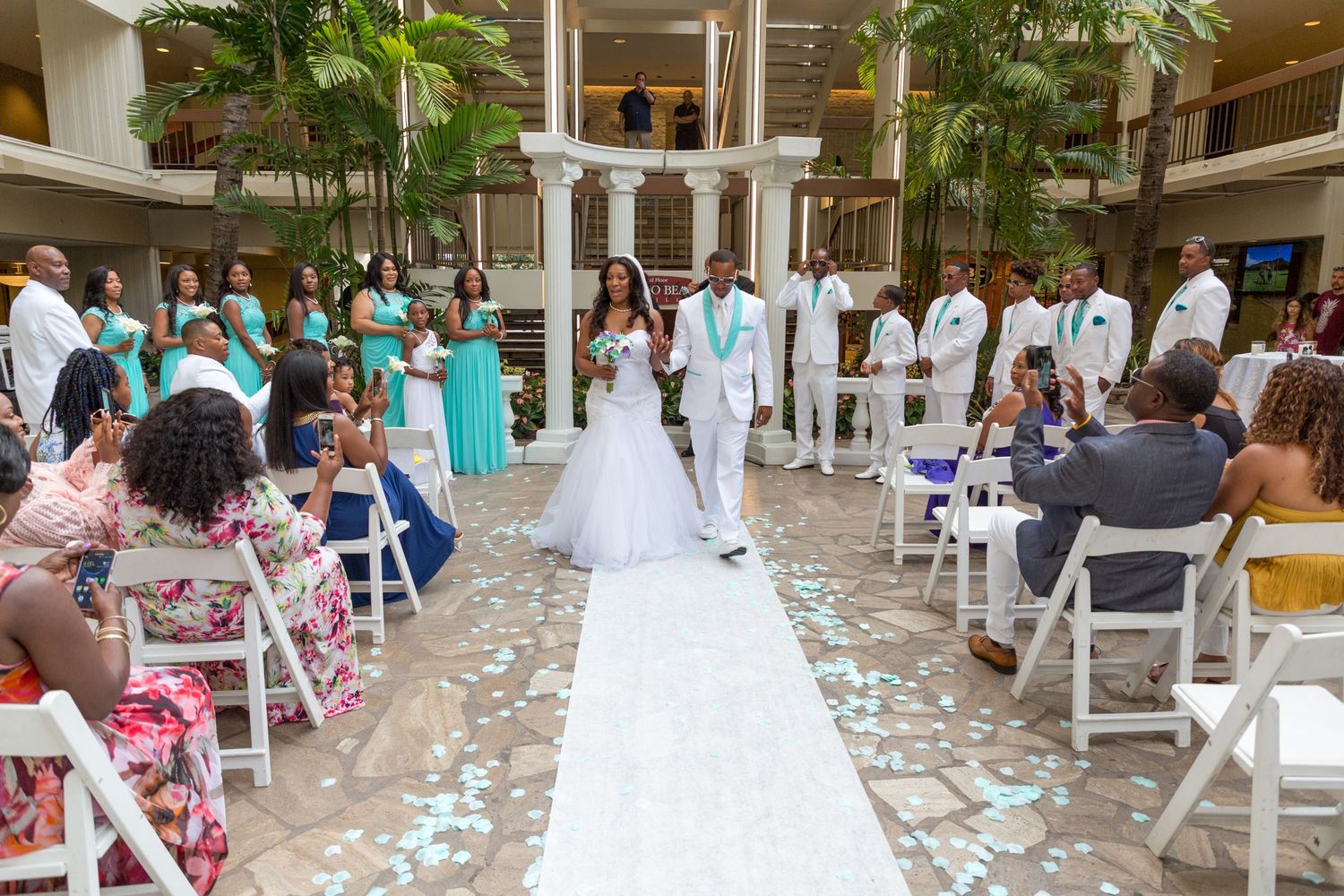 Wedding ceremony in the tropical courtyard of the Hilton Waikiki Beach hotel of Honolulu with white chairs and teal-dressed attendees.