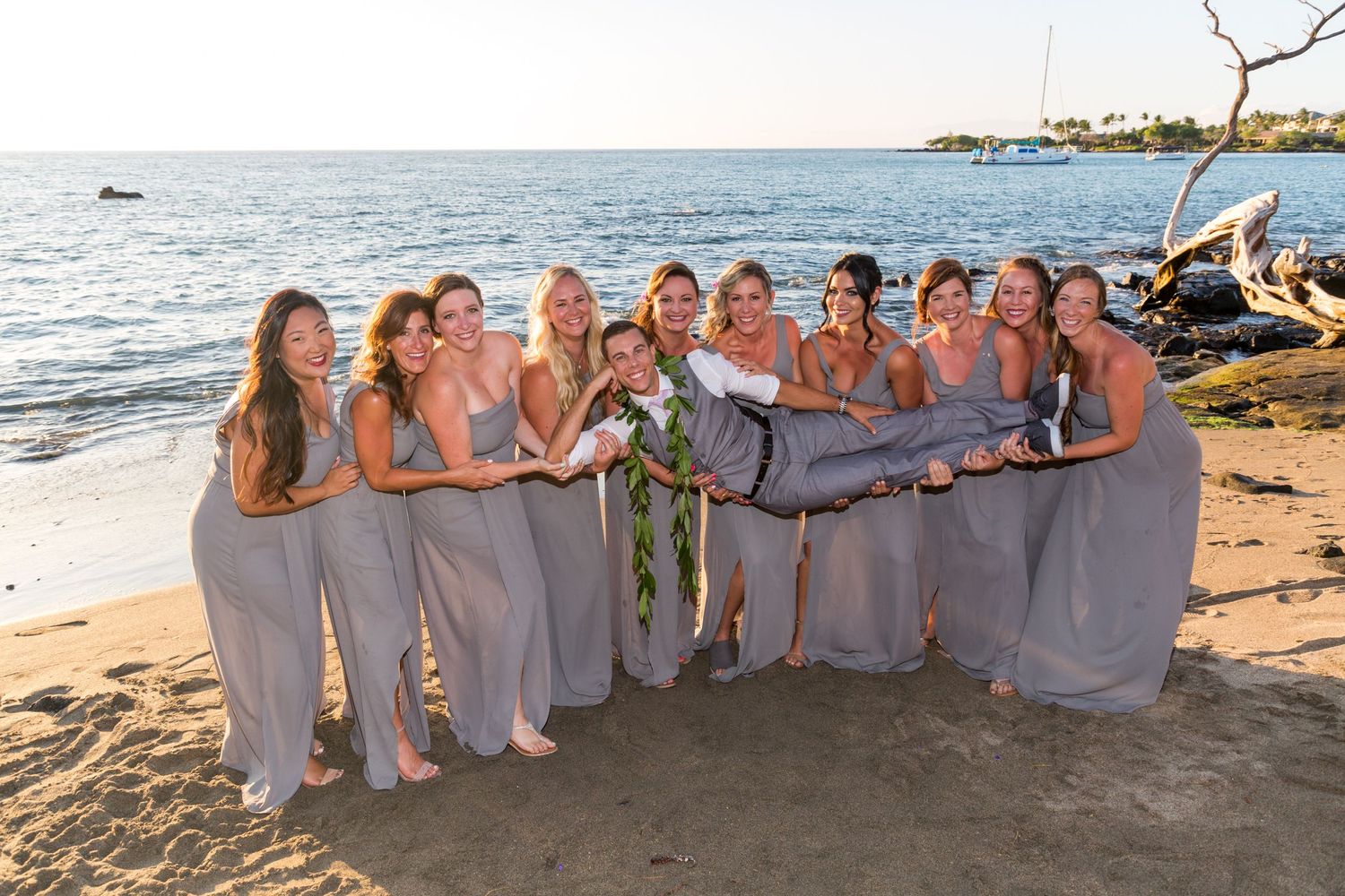 A group wearing matching lavender dresses poses together on a tropical beach at sunset during a wedding celebration.