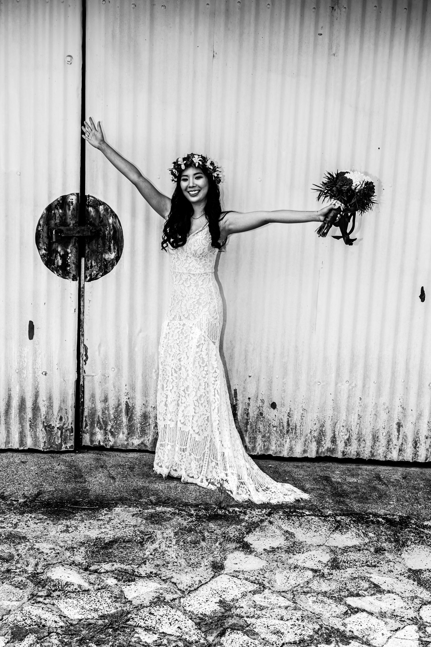 A joyful bride in a lace wedding dress poses against a rustic wall at Anna Ranch Heritage Center in Waimea, Hawaii.