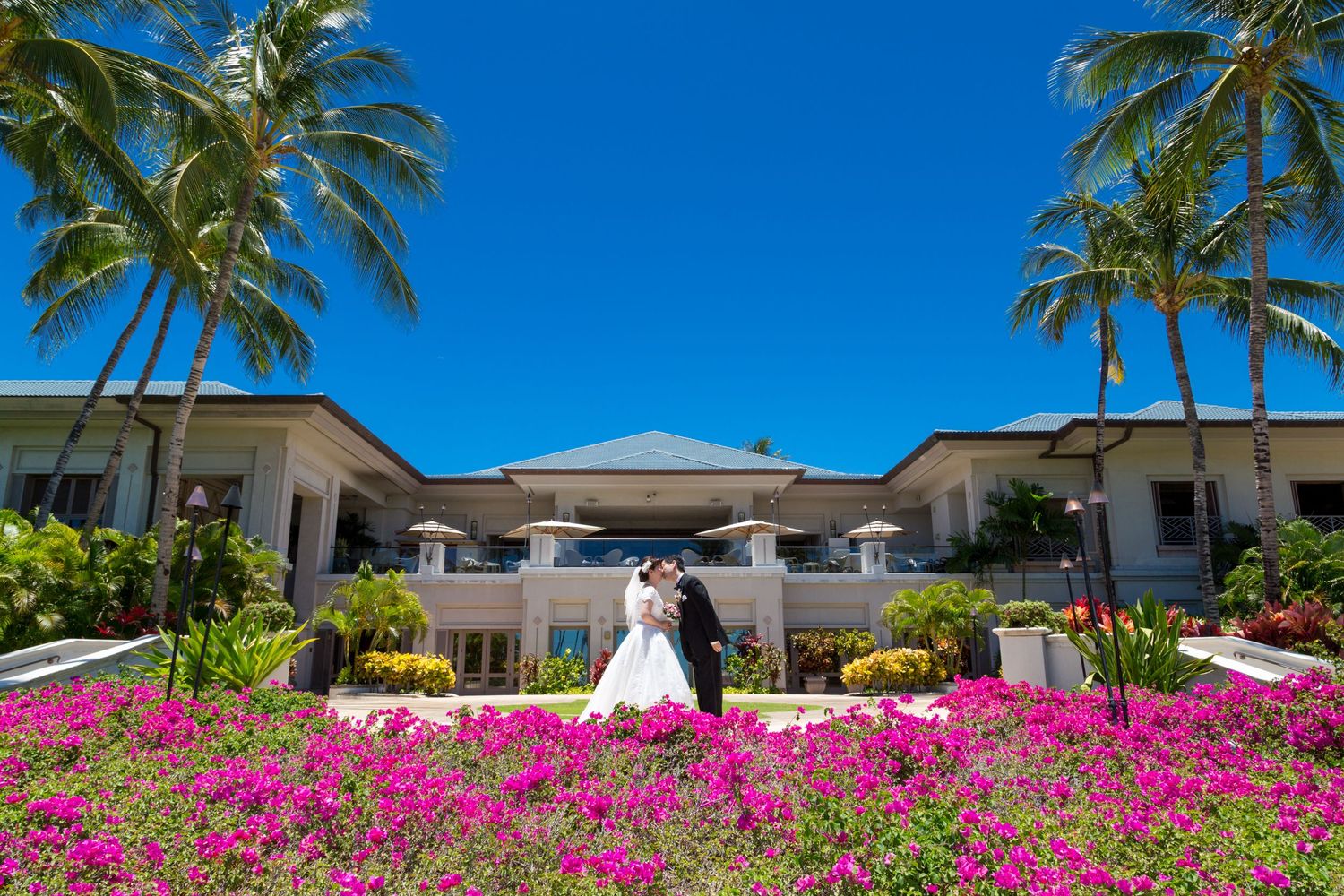 Couple in wedding attire at the Fairmont Orchid hotel in Waikoloa, Hawaii, with vibrant pink flowers in foreground.