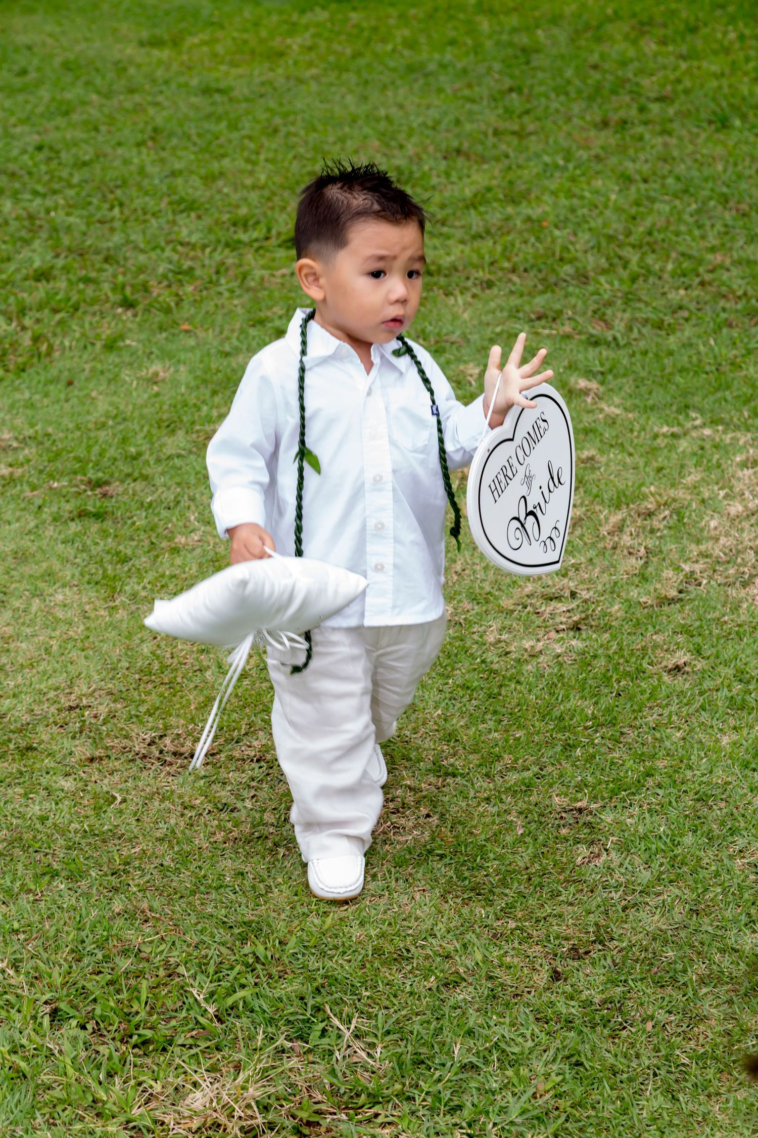 Small child in all white outfit holding a decorative sign while standing in green grass in Honolulu.